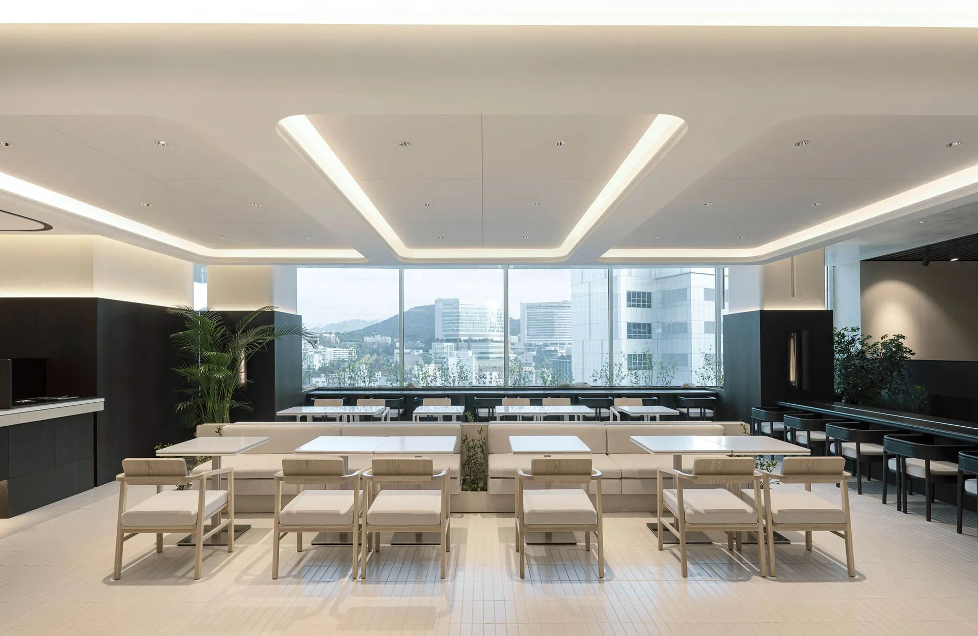 Modern minimalist restaurant interior featuring light wood chairs, white upholstered banquettes, white tables, and large panoramic windows overlooking a cityscape and mountains.