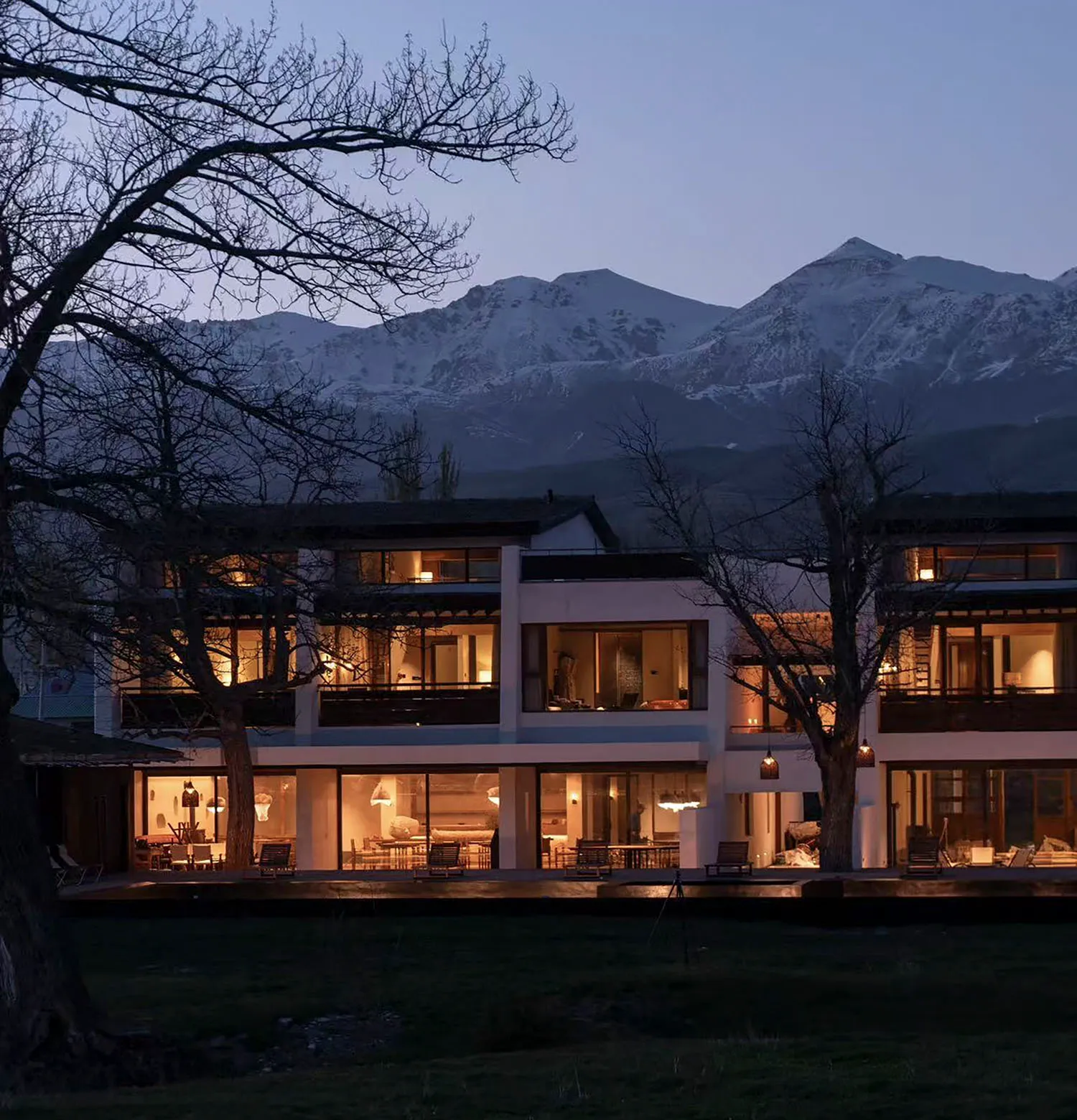 A contemporary two-story villa with large glass windows and wooden details, illuminated from within at twilight, set against snow-capped mountains with bare trees in the foreground.
