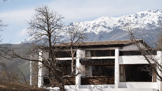 Modern white stucco mountain villa with wooden details, set against a dramatic backdrop of snow-covered mountains in winter.
