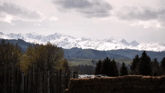 遠處白雪覆蓋的山峰,從鄉村田野望去,前景有木柵欄與乾草堆,頭頂是多雲的天空。