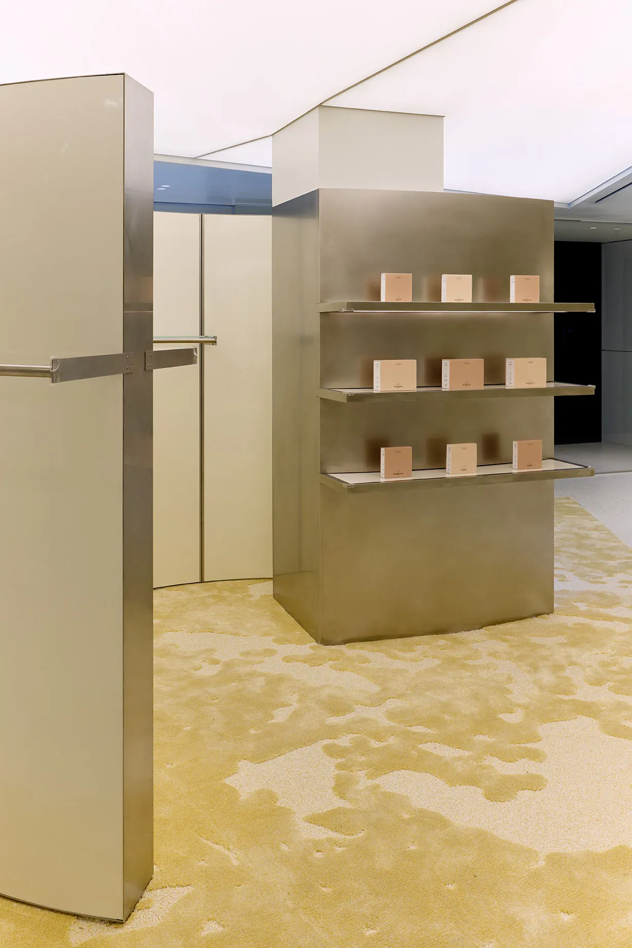 Modern minimalist retail interior featuring a tall brushed stainless steel display shelving unit with integrated lighting, set on a thick, abstract-patterned chartreuse and cream carpet.