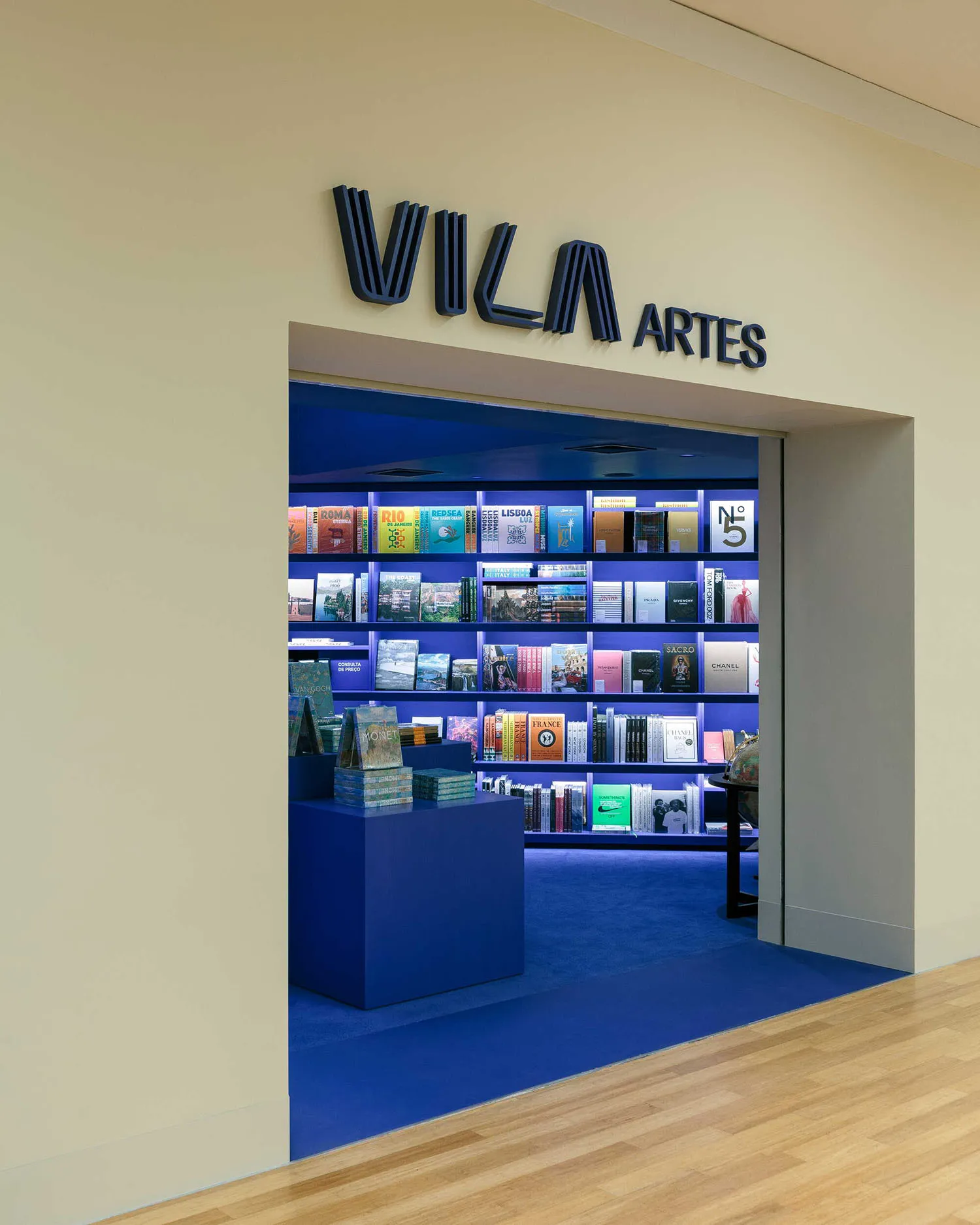 Entrance view of a modern bookstore with intense blue interior lighting, bookshelves, and a blue display platform, featuring a dark blue 'VILA ARTES' sign above the doorway.