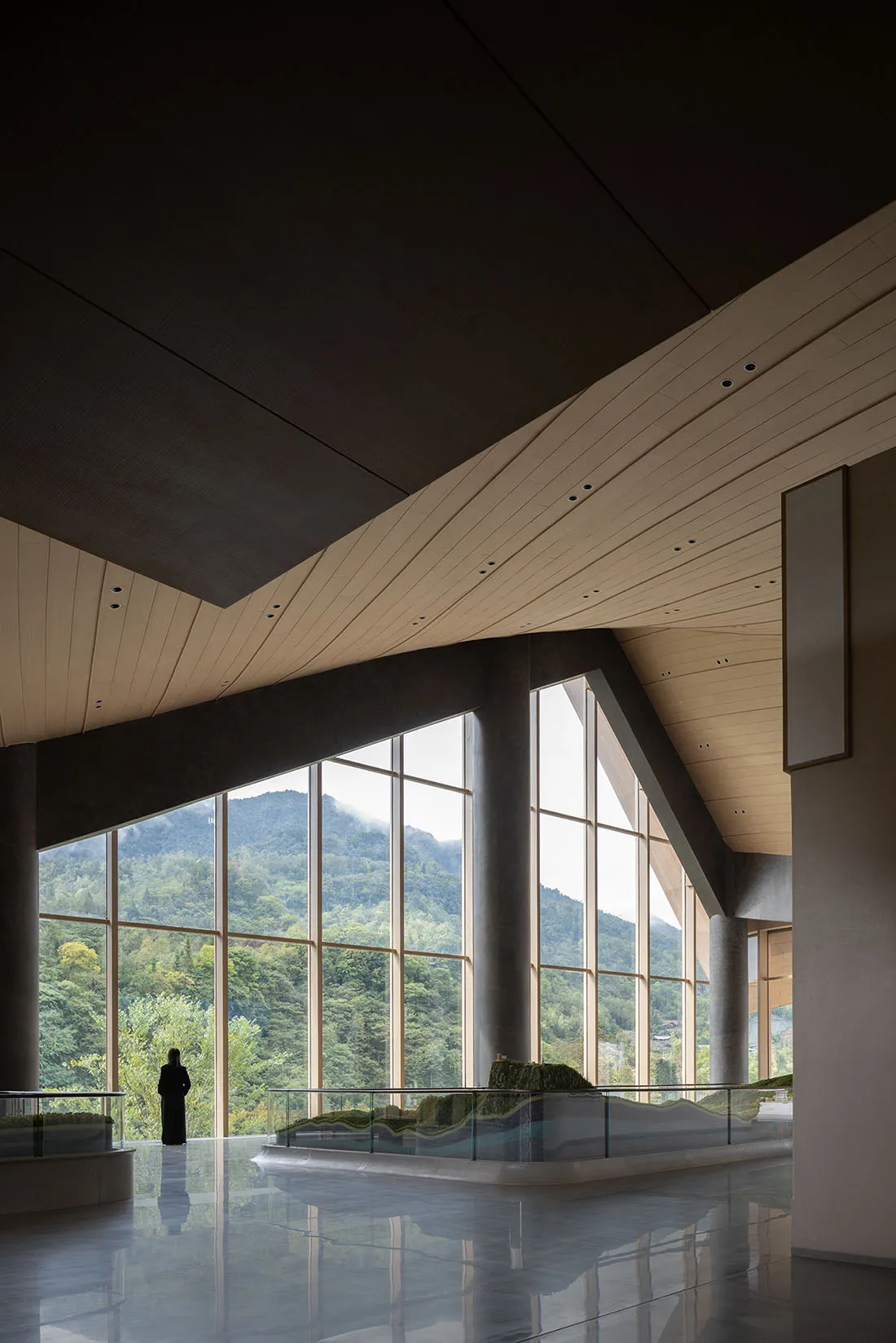 Spacious modern lobby interior with polished concrete floor, curved wooden ceiling, massive floor-to-ceiling windows overlooking green mountains, and a person standing near the glass.