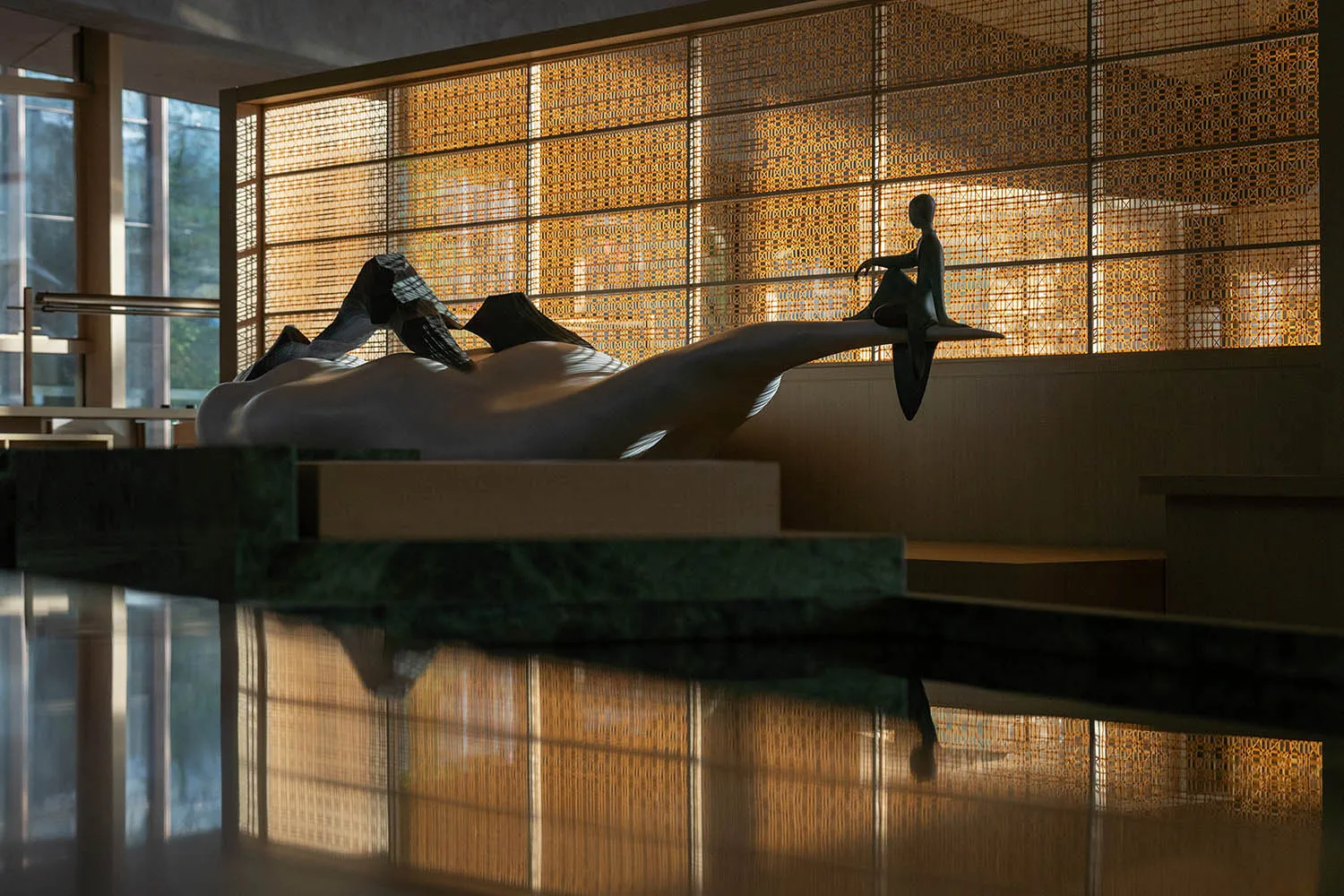 Modern abstract white marble sculpture with a bronze seated figure displayed on a green marble platform in a sunlit contemporary lobby with woven wooden lattice screens.