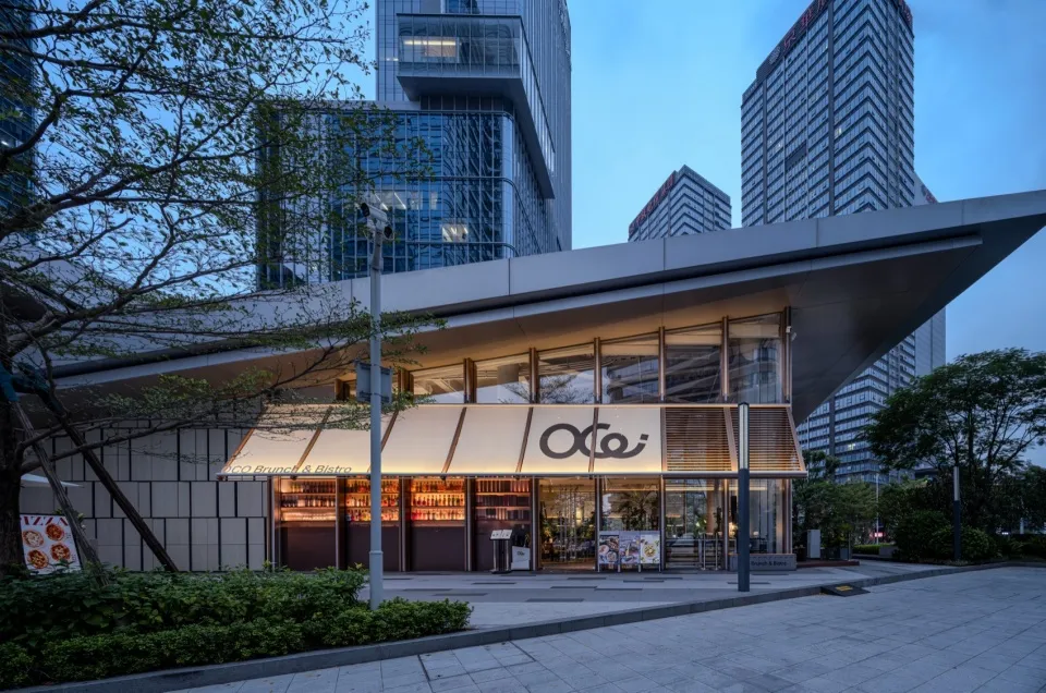 Evening view of OCO Brunch & Bistro modern glass and concrete facade with illuminated signage and warm interior lighting, surrounded by high-rise buildings in a city setting.