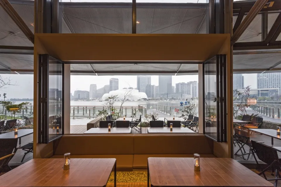 現代餐廳室內空間,木製餐桌、固定沙發座椅與大型可折疊玻璃門,面向水岸城市景觀,遠處可見特色白色建築。