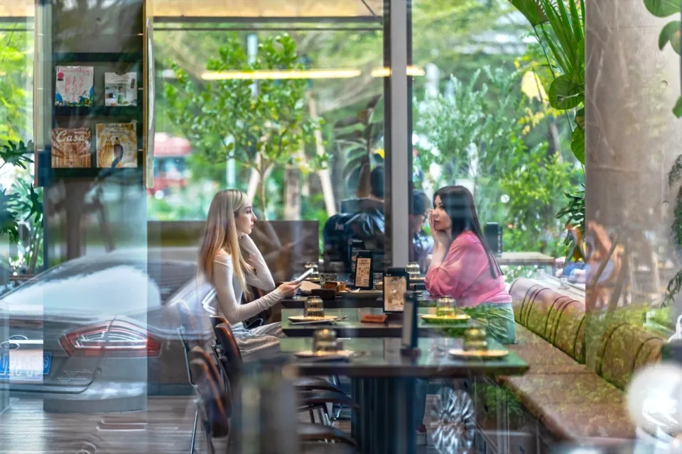 Two women sitting at a table inside a modern glass-walled restaurant overlooking a lush green garden, with marble bench seating and warm wooden tones.