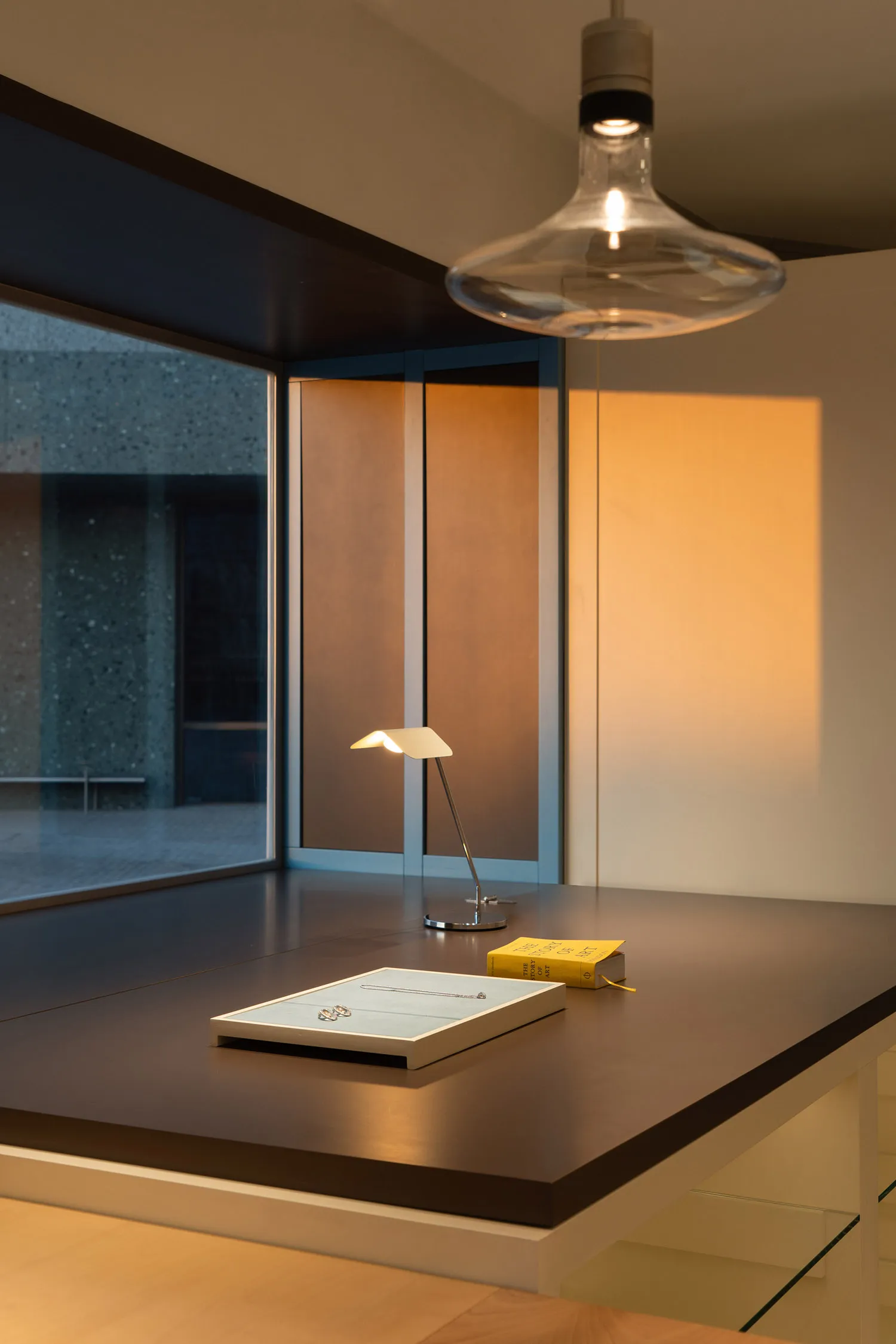 Modern minimalist study corner featuring a dark walnut desk, a lit glass pendant lamp, a small white task lamp, and two books on a light blue tray with jewelry.