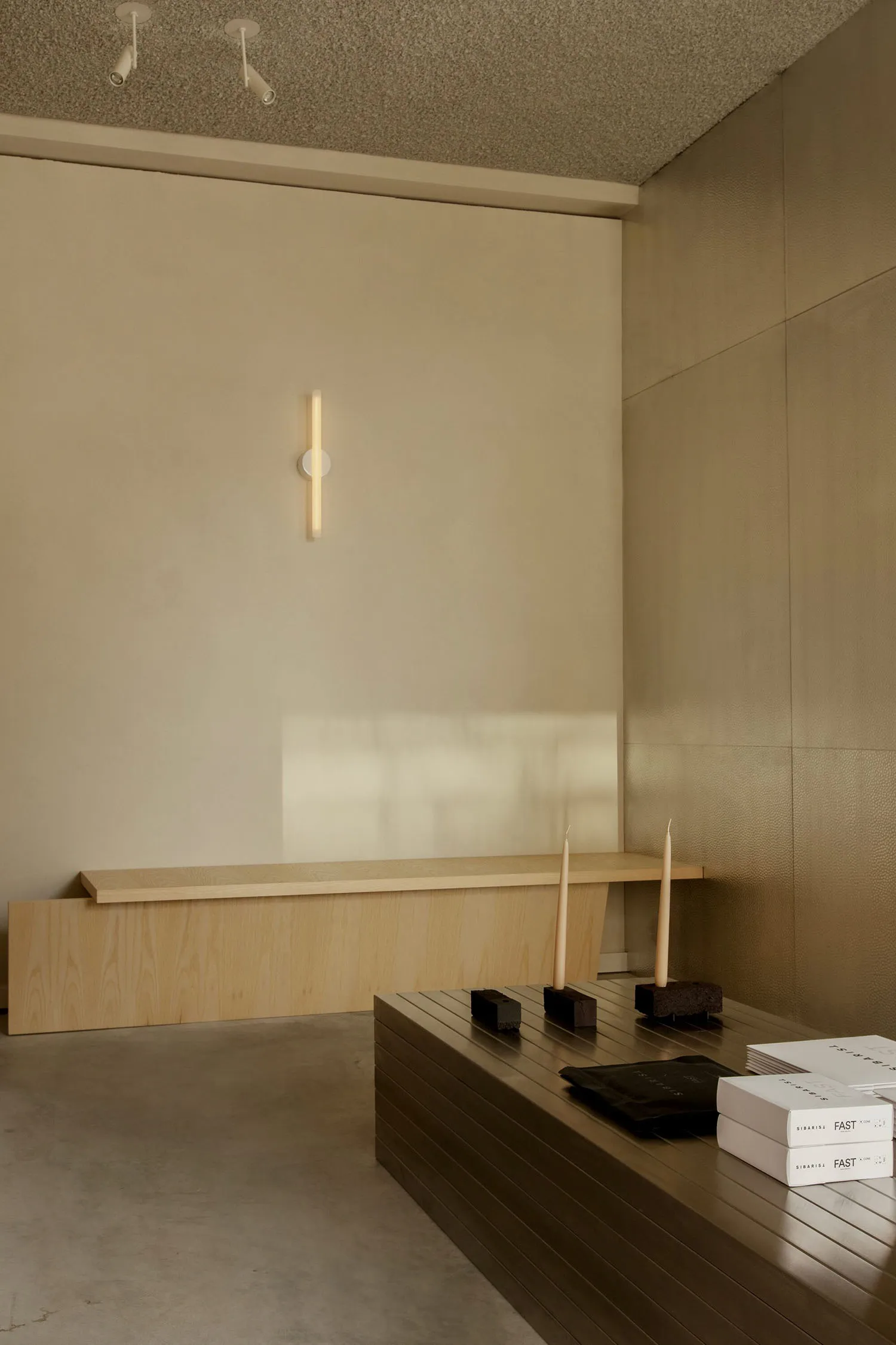 Minimalist living room corner featuring a light oak floating console, dark wood coffee table, textured beige walls, and vertical wall sconce lighting.