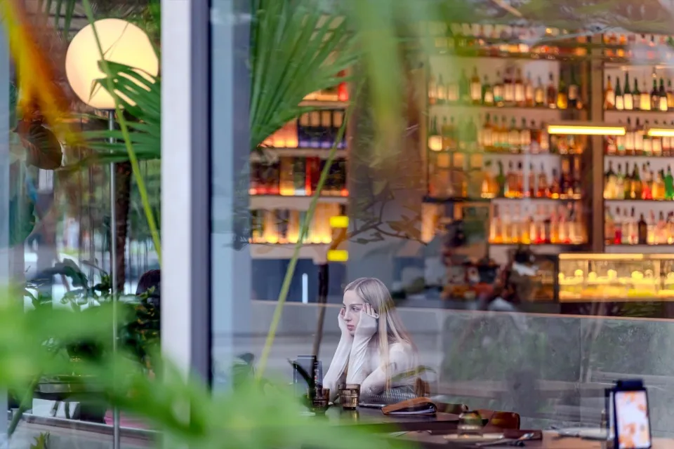 A young woman with long blonde hair sitting at a modern restaurant bar counter, viewed through large glass windows and tropical plants, with illuminated backlit bottle shelves in the background.