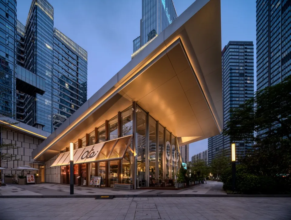 Evening view of a modern minimalist cafe with large glass windows, cantilevered concrete roof with warm lighting, and surrounding high-rise buildings in a city plaza.