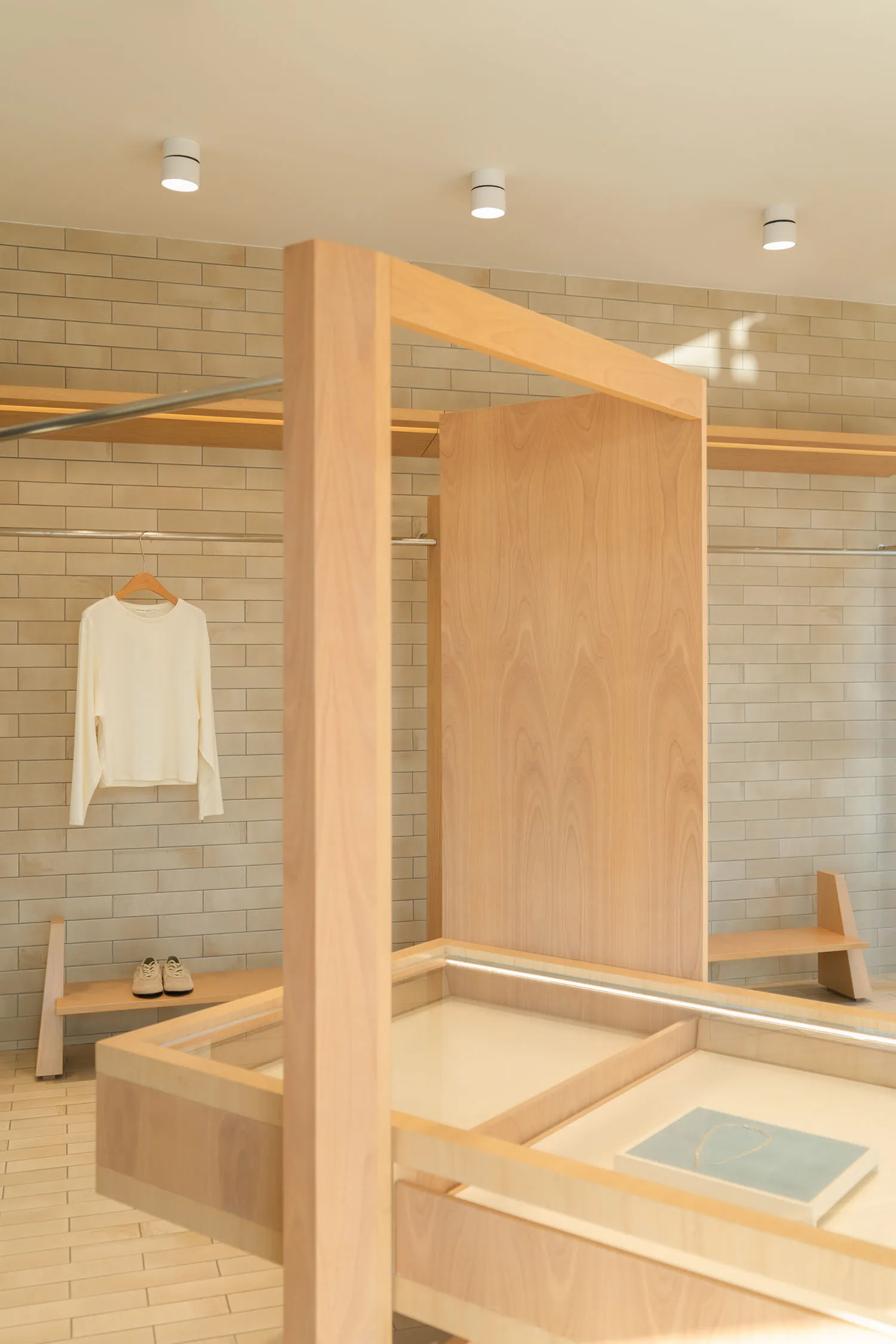 Modern minimalist retail interior with light oak wood display counters, shelving systems, and beige brick walls, featuring a white long-sleeve shirt hanging on a clothing rack and a jewelry display tray.