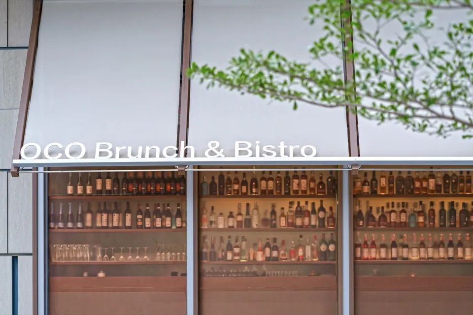 現代餐廳酒吧展示,玻璃窗後方有大量酒瓶陳列架、玻璃杯具與溫暖燈光,上方有OCO Brunch & Bistro招牌。