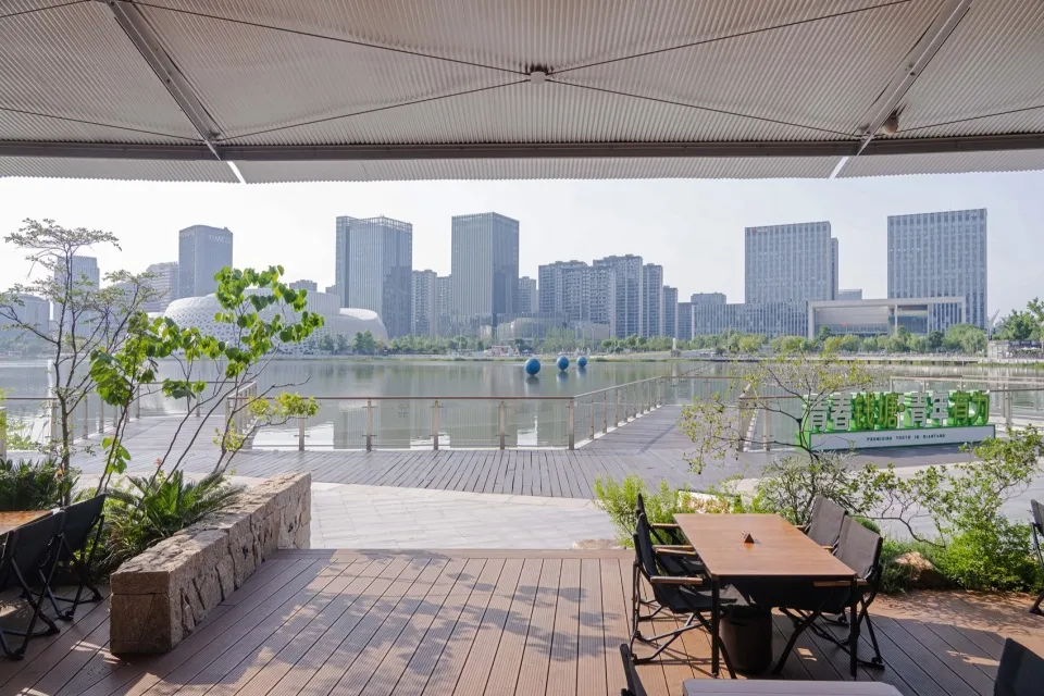 Modern wooden terrace with dining table and chairs overlooking a lake and the Songshan district city skyline in Taipei, with lush greenery in the foreground.