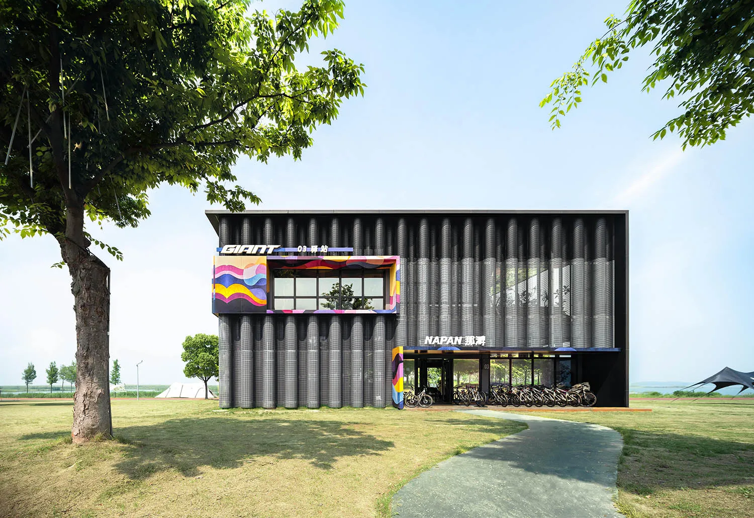 Modern black corrugated metal bicycle hub building with colorful graphic panels and numerous bicycles parked under the canopy on a sunny day.