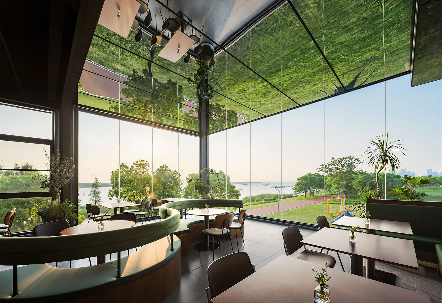 Modern restaurant interior featuring curved green velvet banquettes, wooden tables, and expansive floor-to-ceiling windows overlooking a lake and lush parkland.