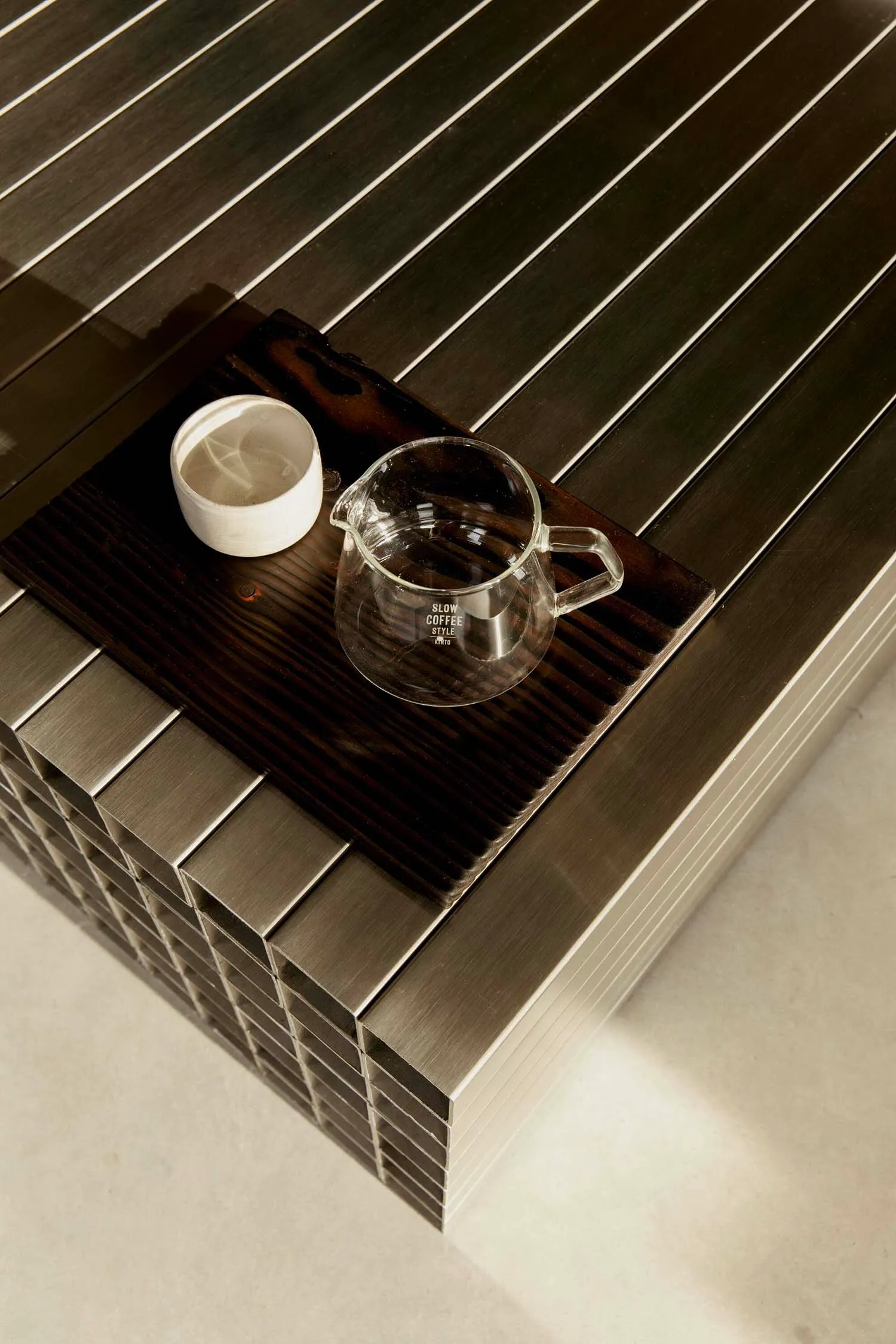 Close-up top view of a brushed stainless steel coffee table featuring a dark wooden tray holding a glass coffee server and a white ceramic cup.