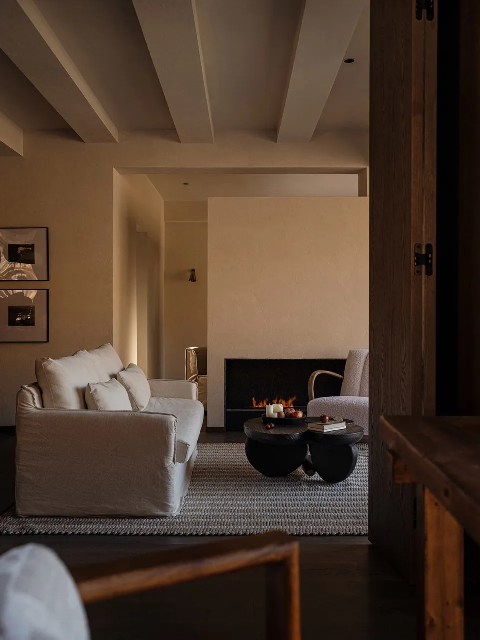 Modern minimalist living room featuring a white linen sofa, sculptural black coffee table, lit fireplace, and textured beige plaster walls with exposed wooden ceiling beams.