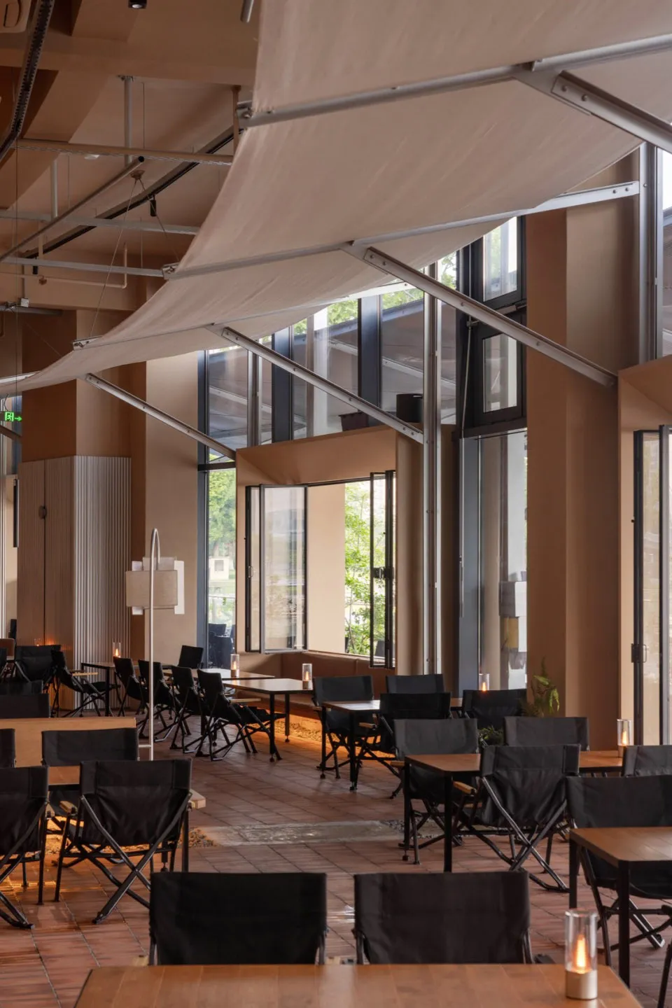 Modern restaurant interior featuring terracotta floor tiles, black folding chairs, wooden tables, large windows with greenery views, and a large suspended white canvas canopy structure.