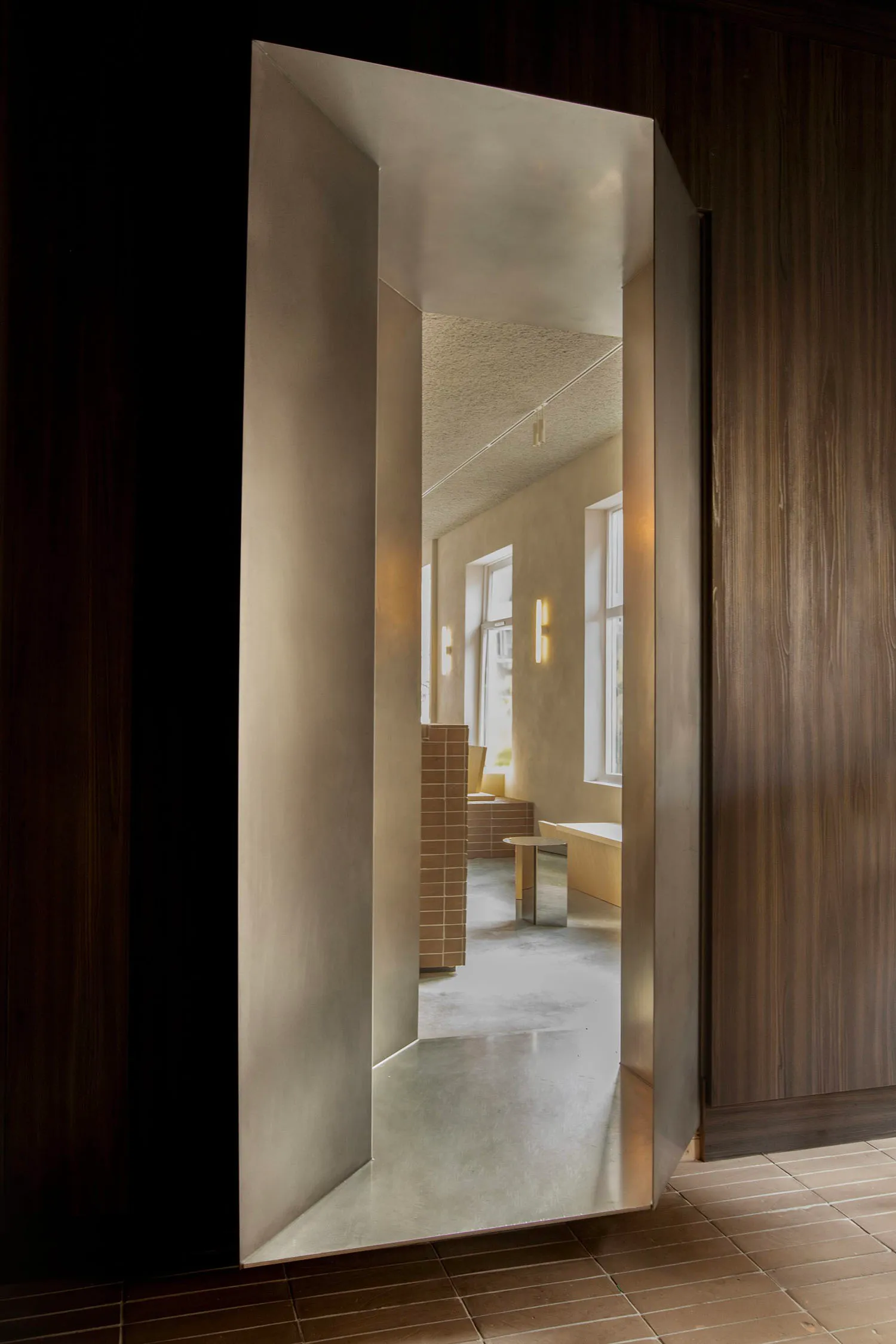 View through a thick concrete doorway into a minimalist hallway featuring raw concrete surfaces, warm wood paneling, terracotta tile accents, and natural daylight.