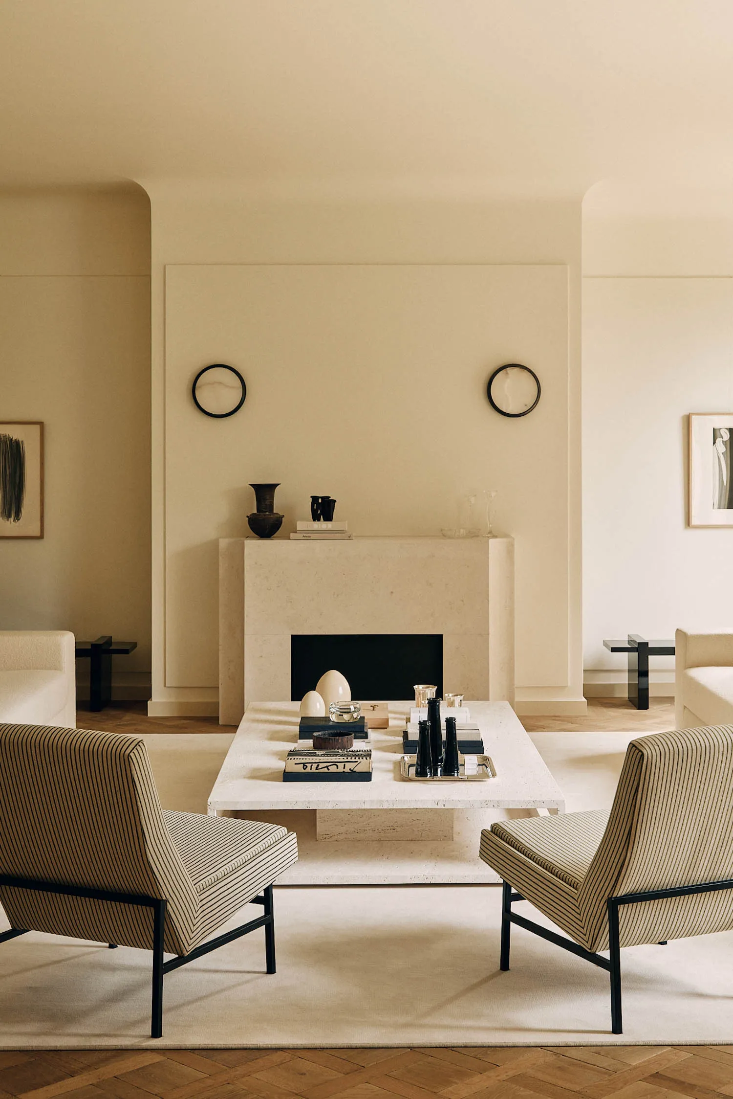 Modern minimalist living room featuring a large travertine fireplace, two striped upholstered armchairs, a matching travertine coffee table, and neutral beige walls with decorative objects.