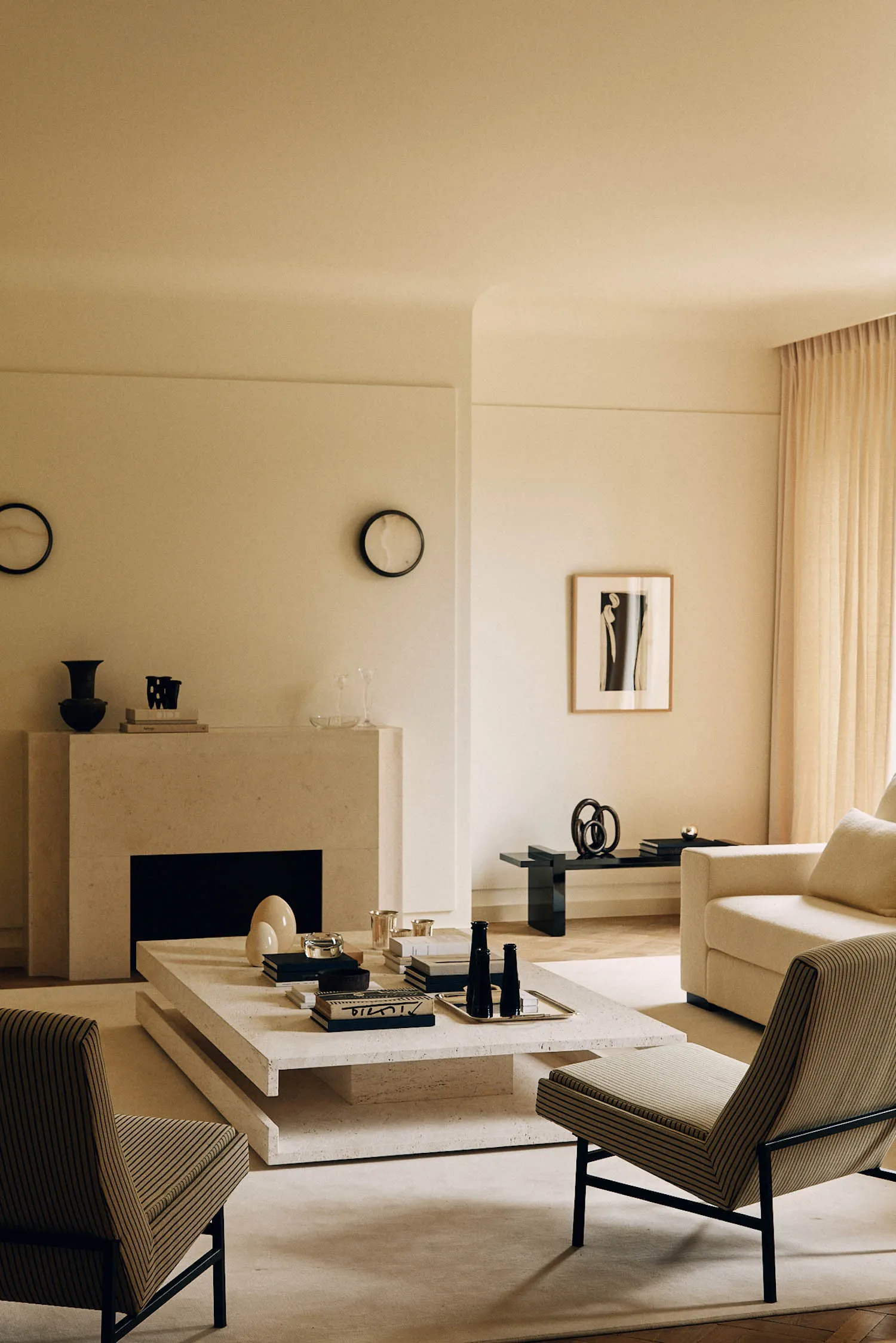 Modern minimalist living room featuring a large travertine coffee table, matching fireplace mantel, two striped armchairs with black metal frames, a cream sofa, and warm ambient lighting.