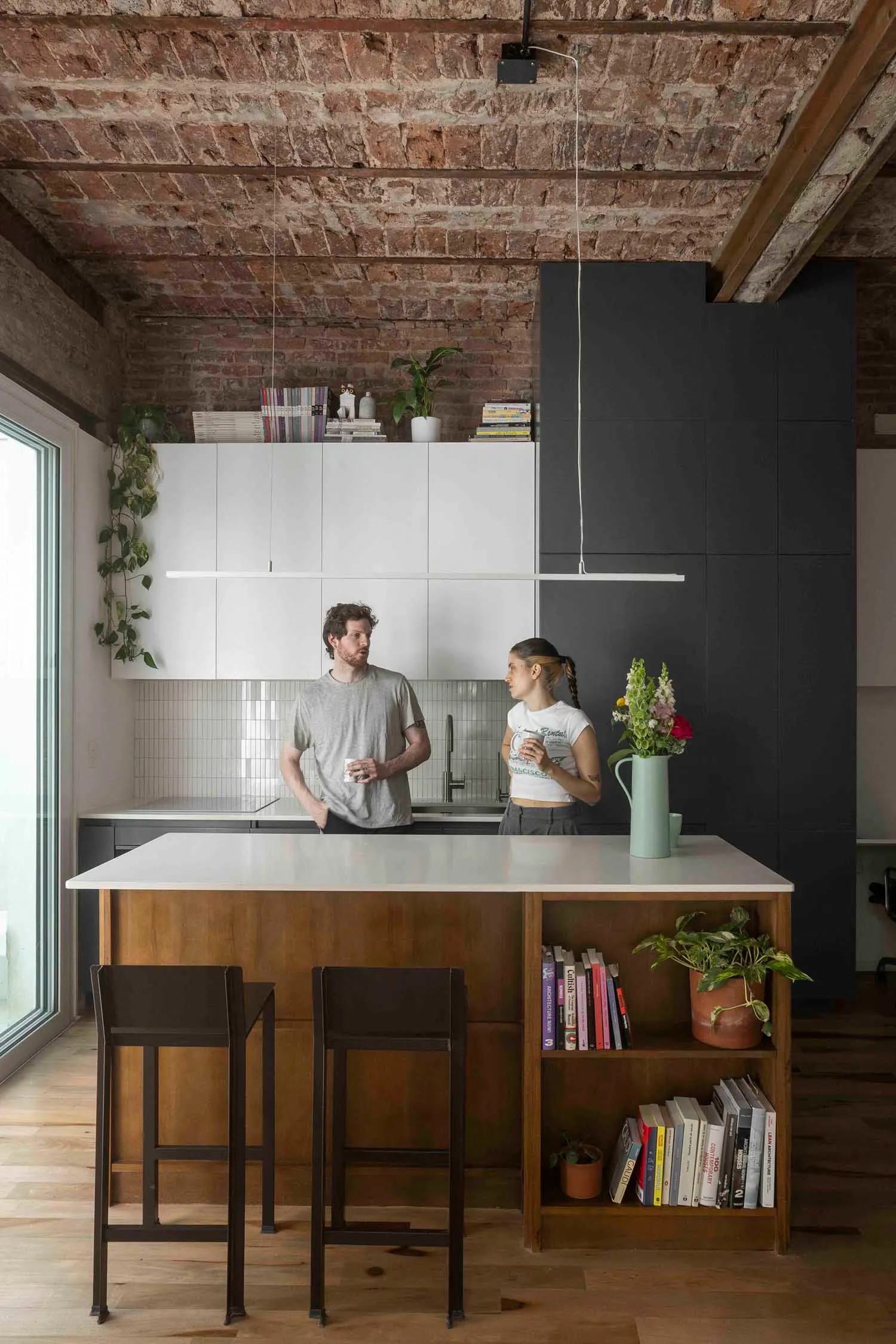 Modern industrial loft kitchen featuring a white countertop island with wooden base and open bookshelf, dark grey cabinetry, white upper cabinets, exposed brick ceiling, and two people standing at the counter.