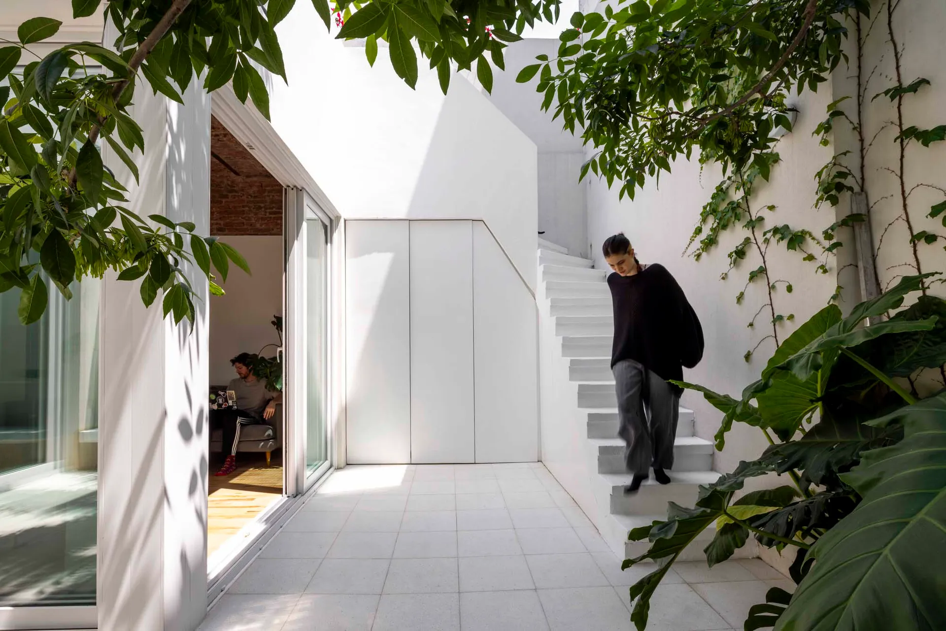 A bright white modern courtyard with a floating staircase on the right, lush green plants and trees framing the scene, and a living area visible through large glass doors on the left where a man is sitting.
