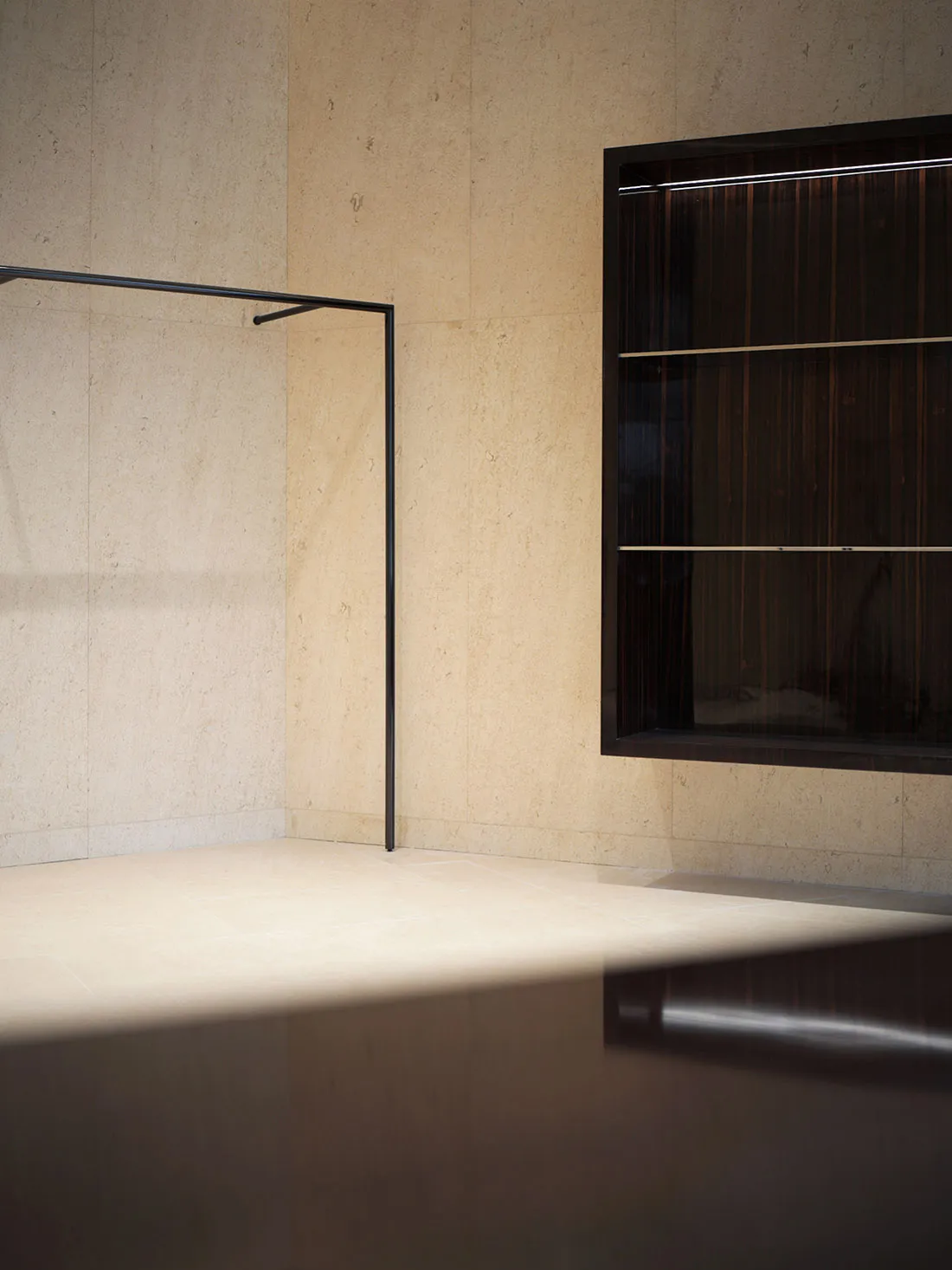 Minimalist bathroom corner featuring large-format travertine walls and floor, a black metal towel rack, and a recessed dark wood display niche with integrated lighting.