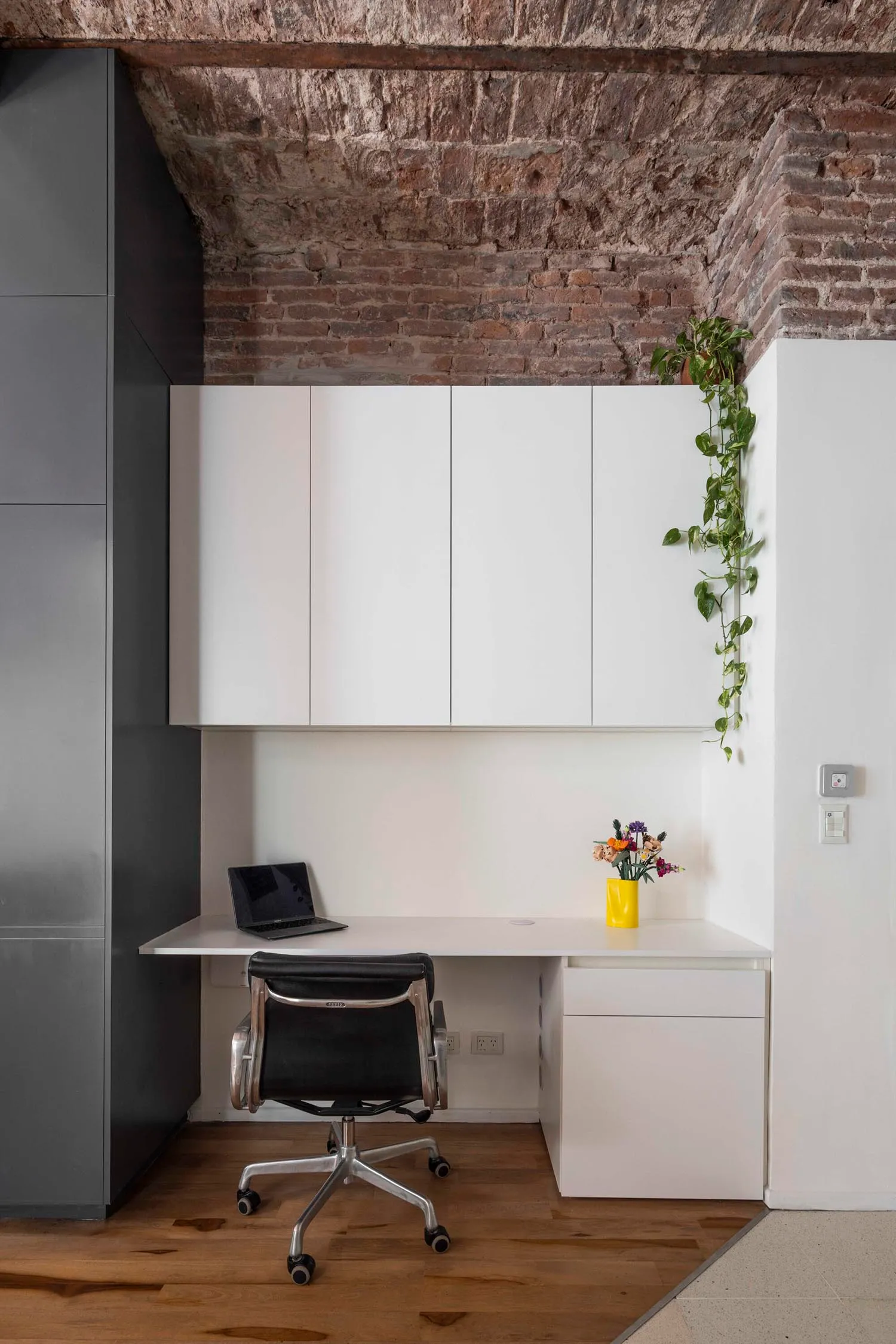 Modern home office nook featuring a white built-in desk, black leather office chair, exposed brick ceiling, and trailing green plants.