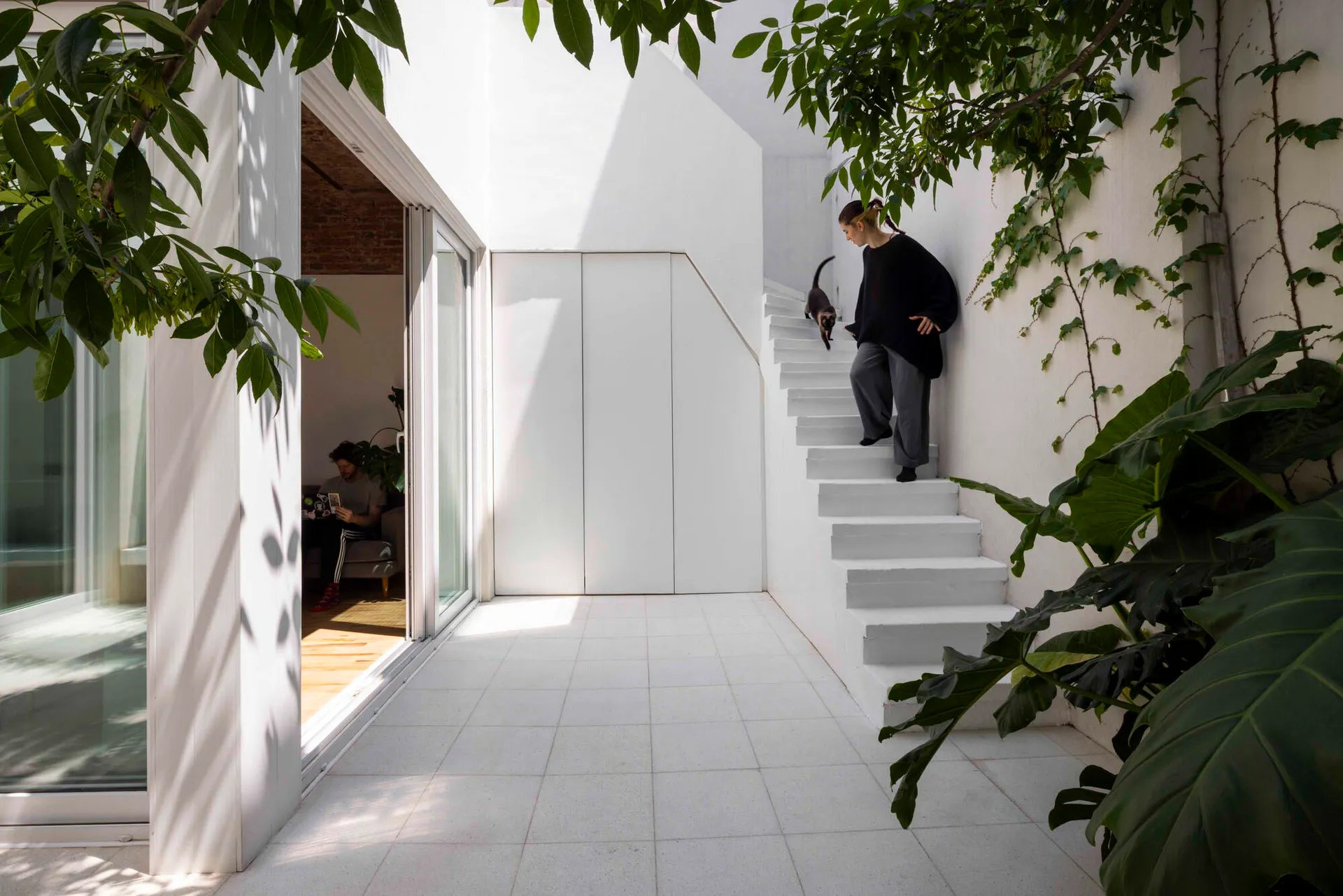 A bright white modern courtyard featuring a floating concrete staircase, large indoor plants, and a woman walking upstairs with a cat, while a man sits in the adjacent living room.