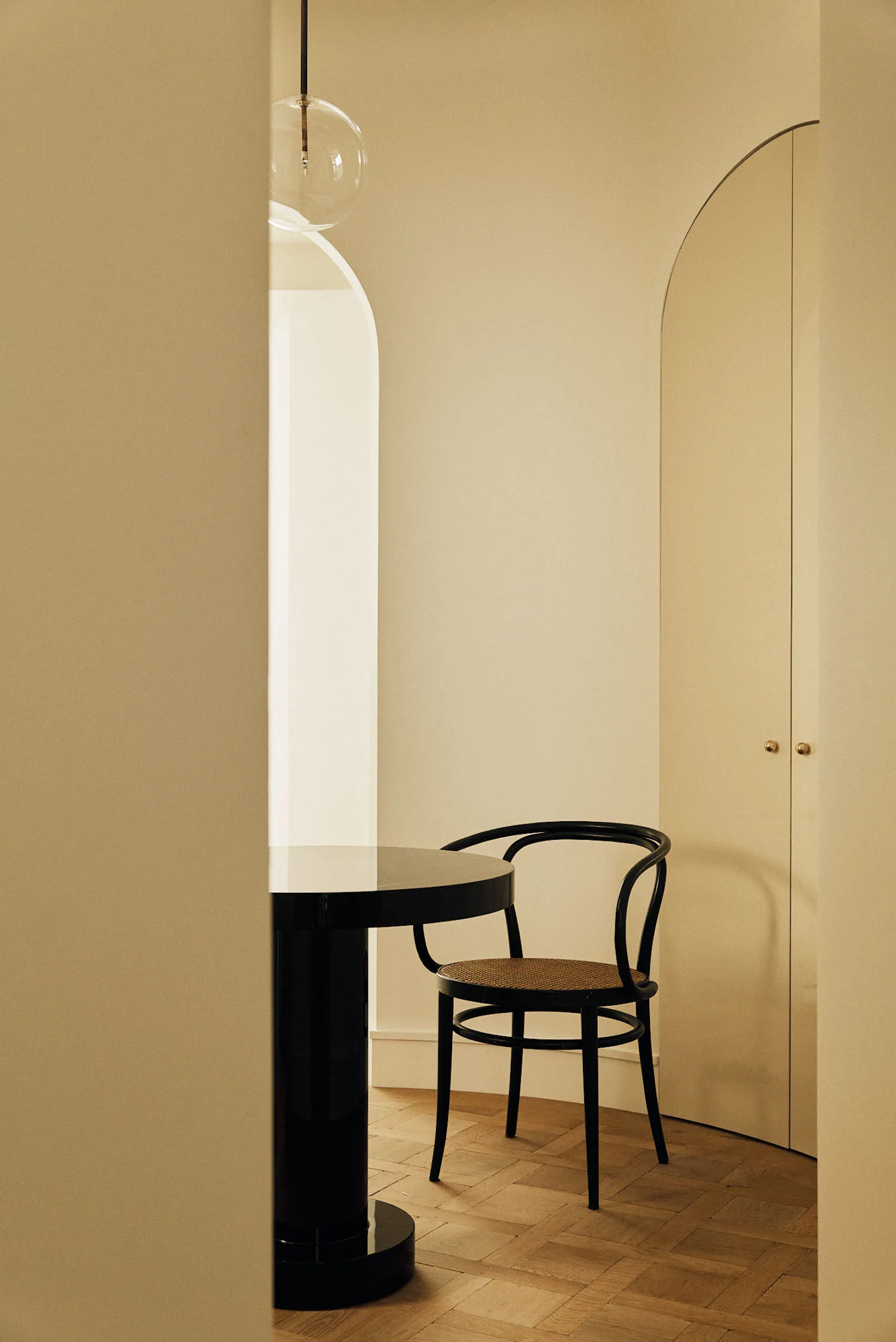 Modern minimalist entryway corner with a black bentwood chair, glossy black round pedestal table, arched doorway, and warm beige walls under a glass globe pendant light.