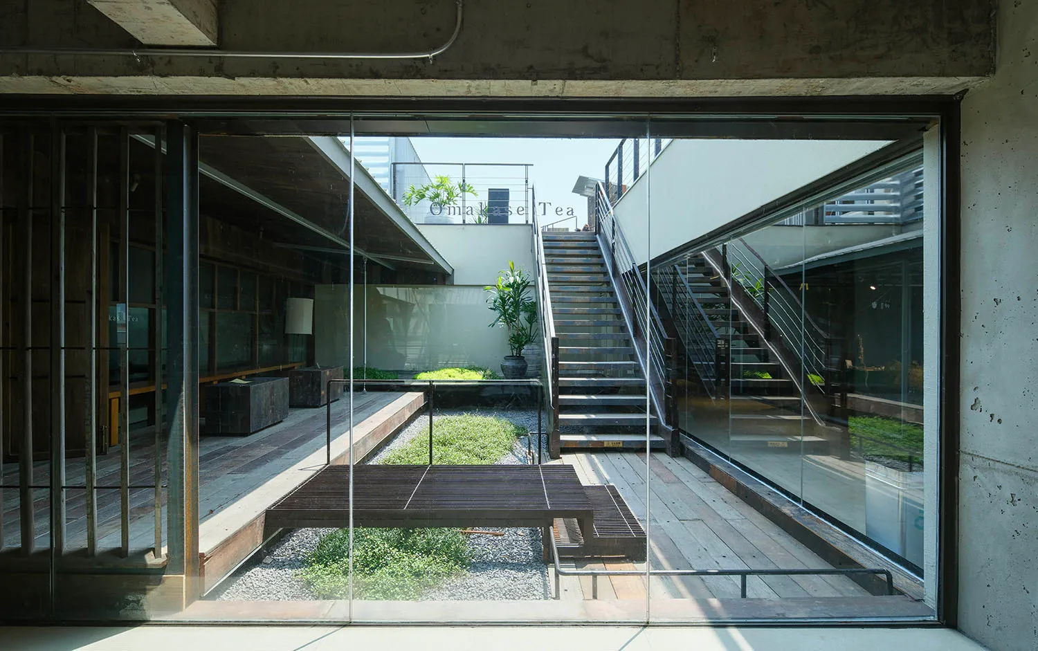 View from inside a modern concrete house looking out onto a sunlit courtyard with wooden deck, gravel garden, potted plants, and external staircase.