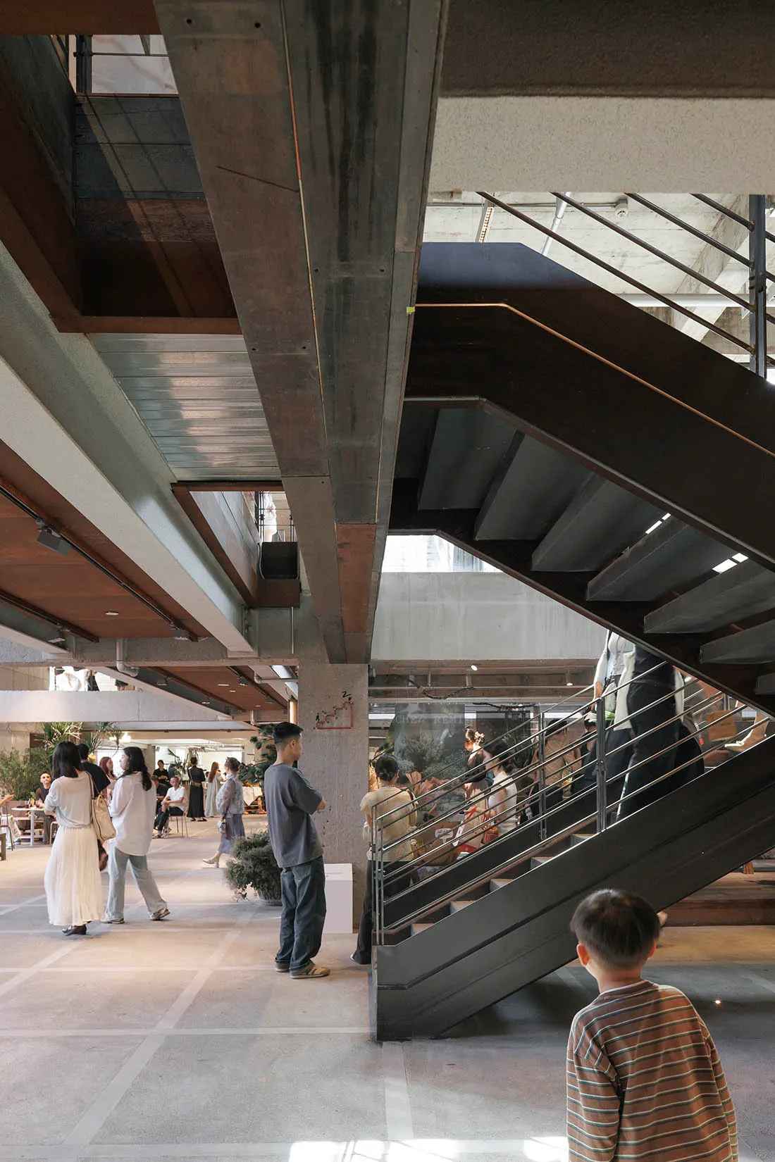People standing in a spacious multi-level brutalist atrium featuring massive exposed concrete beams and a dramatic dark metal staircase.