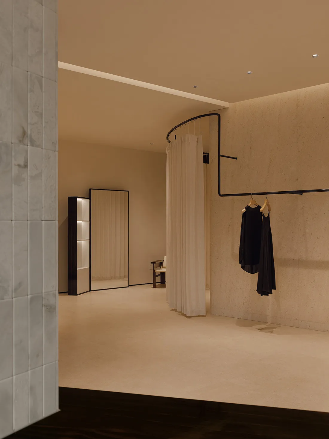 Modern minimalist dressing room featuring raw micro-cement walls, a curved black clothing rail with hanging garments, a tall mirror, and soft recessed lighting.