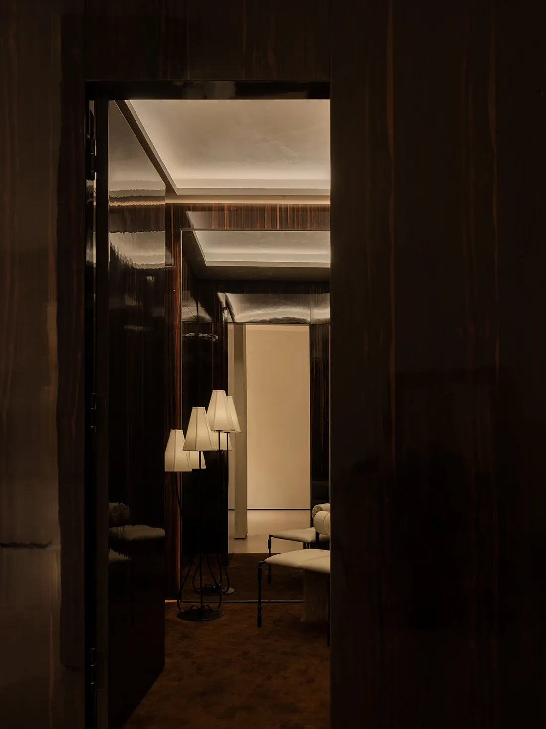 View through a dark macassar ebony doorway into a luxurious hallway featuring multiple sculptural floor lamps with white shades, warm ambient lighting, and rich wood paneling.
