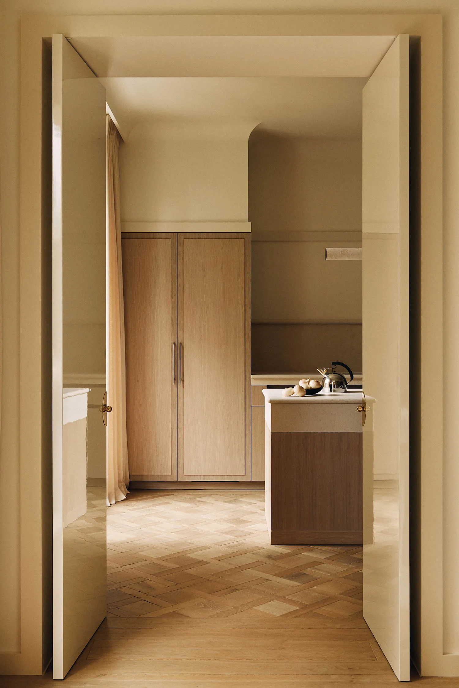 View through open white double doors into a minimalist butler pantry featuring light oak cabinetry, a marble-topped island with a kettle and fruit bowl, and warm herringbone parquet flooring.