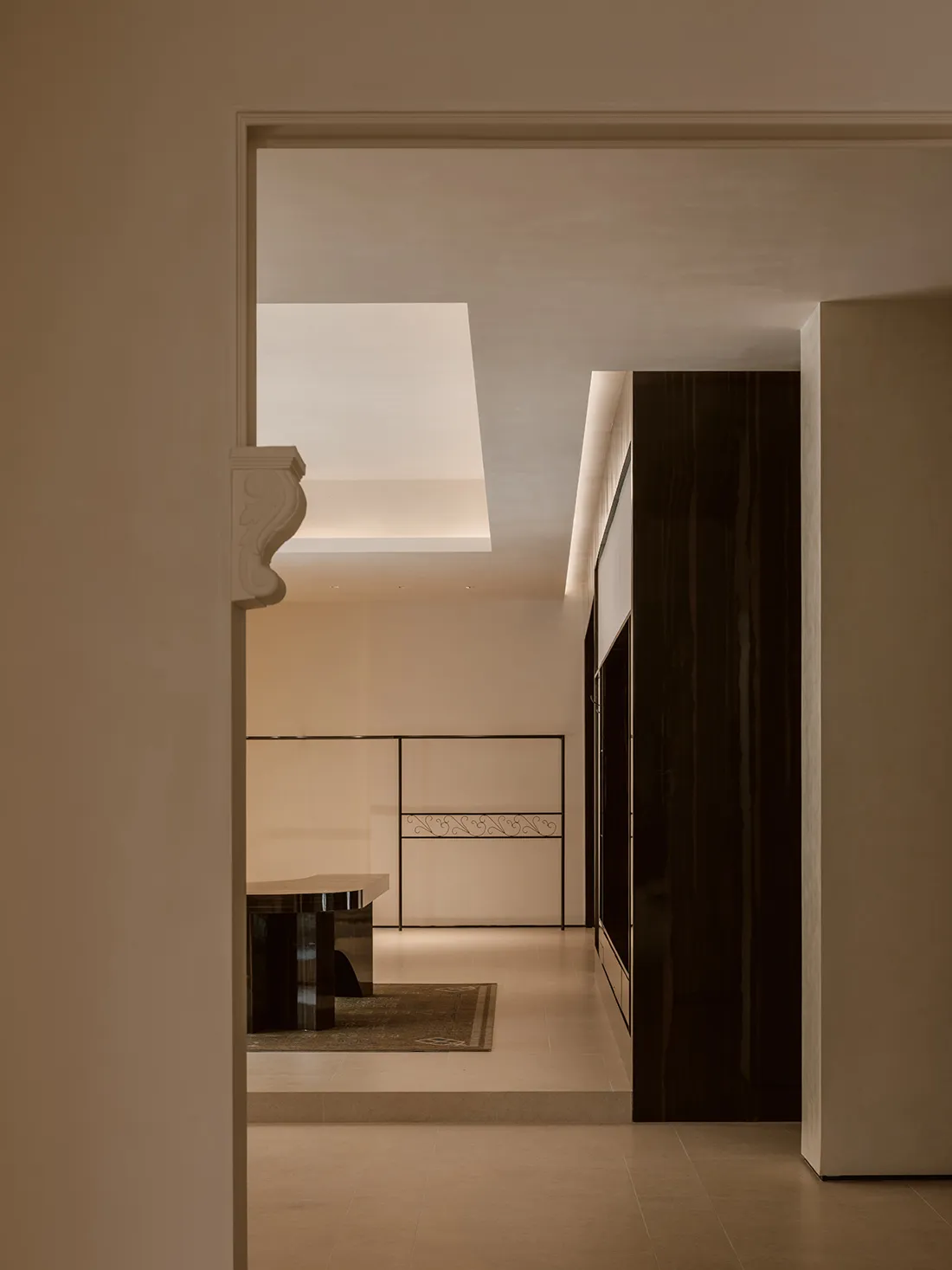 Modern minimalist hallway featuring a large skylight, dark wood cabinetry, light stone flooring, and a decorative metal railing in the background.