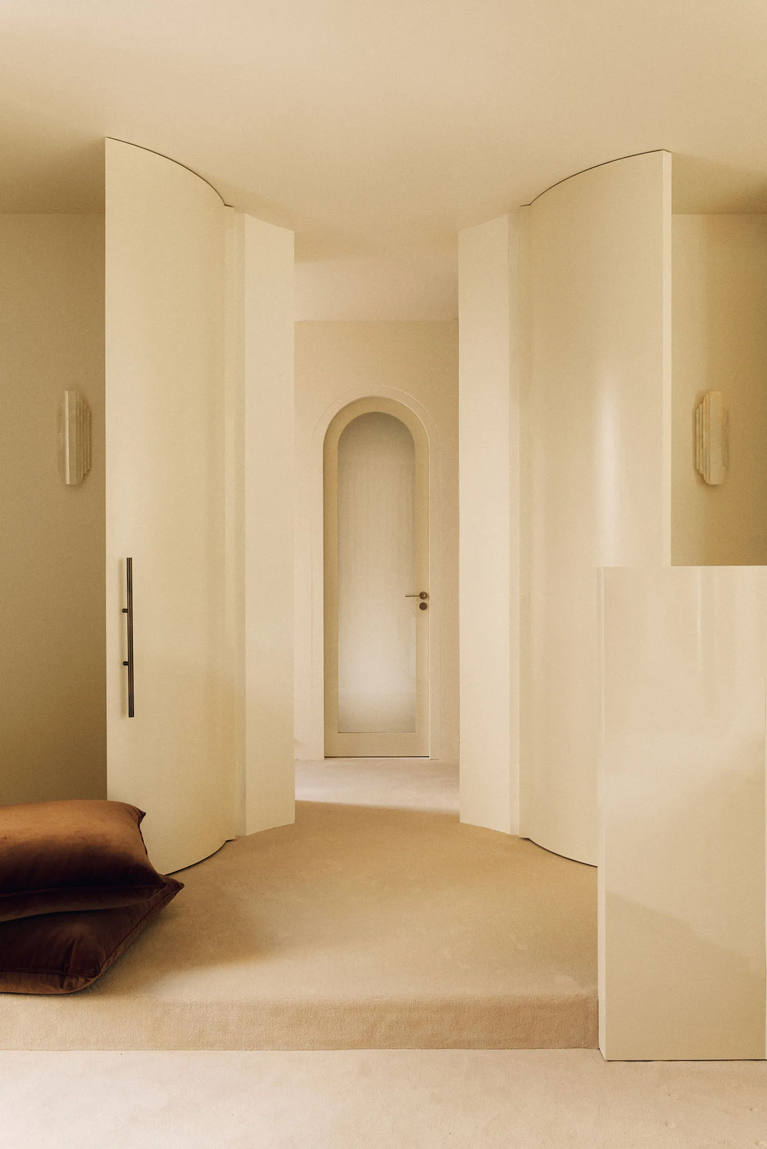Minimalist curved hallway in warm cream tones featuring built-in arched doorway, wall sconces, and two large brown velvet floor cushions in the foreground.