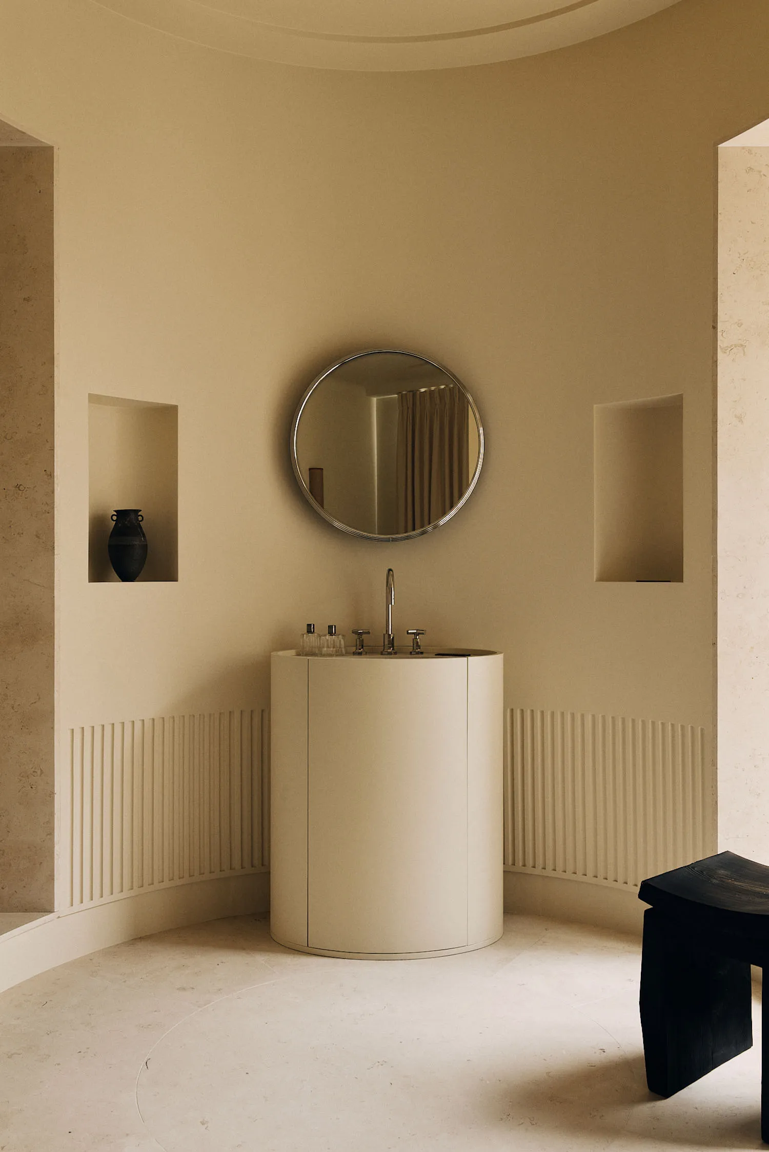 Minimalist powder room with curved micro-cement walls, a cylindrical floating vanity with chrome faucet, round mirror, and dark wooden stool in a warm neutral palette.