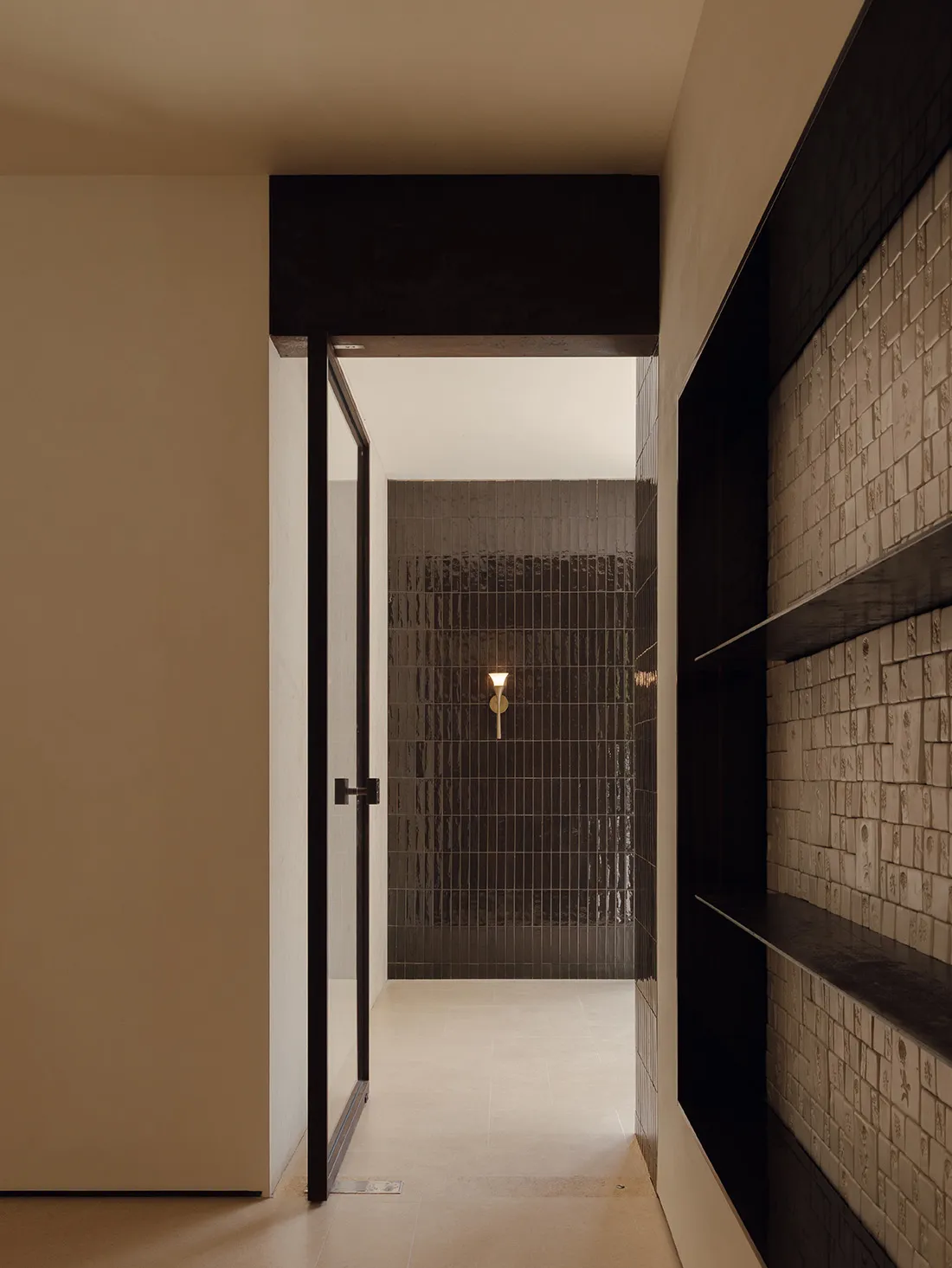 View from a neutral hallway through a black-framed glass door into a shower room lined with dark textured tiles and a brass wall sconce.