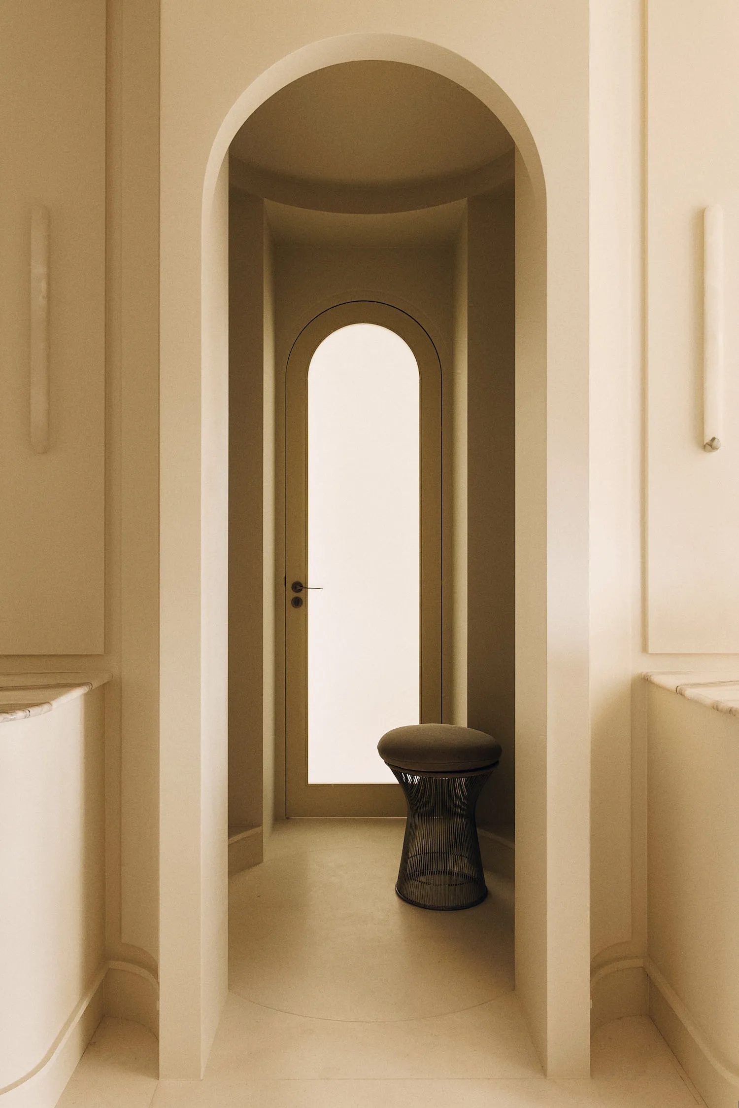 Minimalist entryway featuring consecutive arched niches in warm off-white plaster, with a black wireframe stool placed beneath the inner arch.