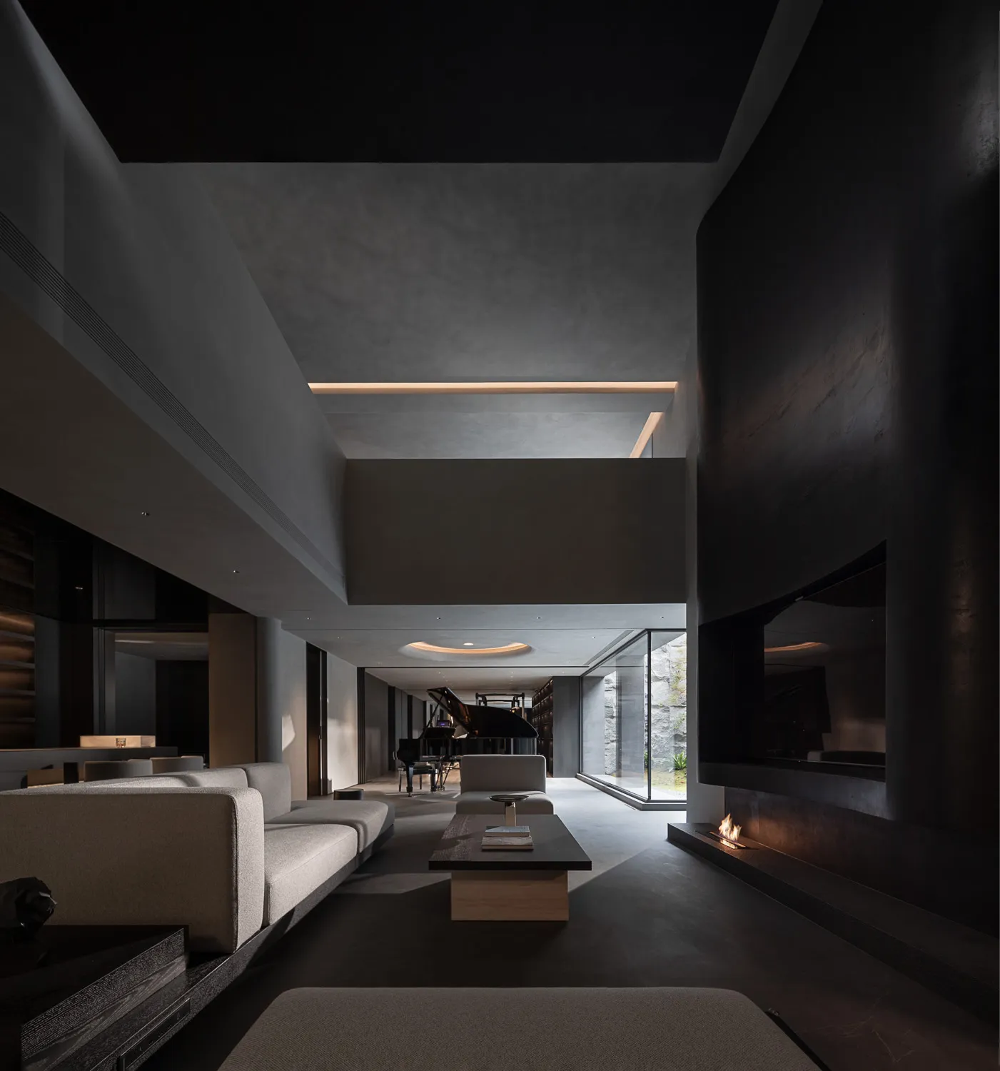 Modern minimalist double-height living room featuring raw concrete walls, a long linear fireplace with visible flame, light grey modular sofas, a dark coffee table, and a grand piano in the distance under dramatic architectural lighting.