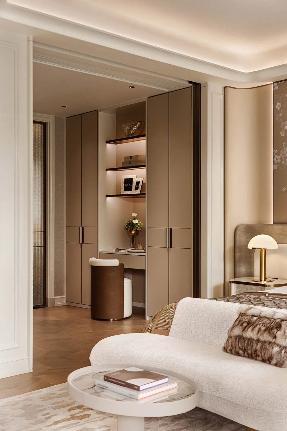 Modern luxury bedroom featuring a built-in illuminated vanity nook with floating shelves, neutral beige cabinetry, a curved white chaise lounge, and marble coffee table.