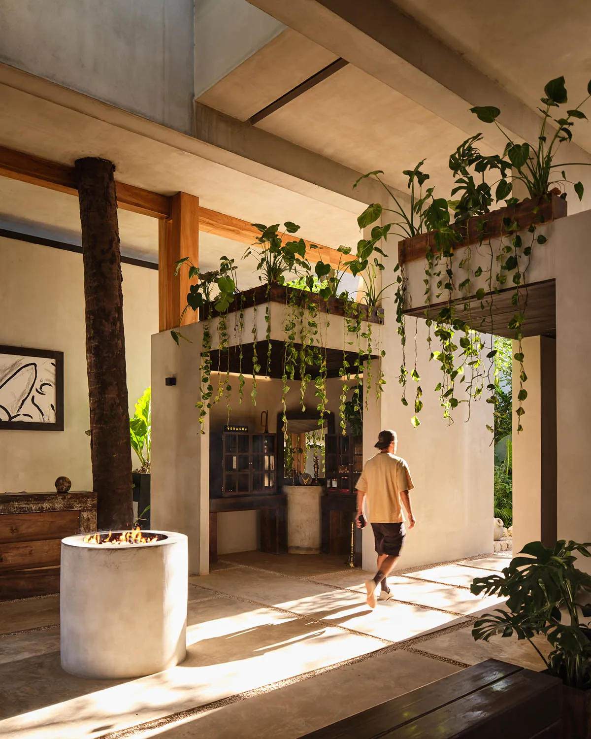 A man walking through a sunlit tropical modern indoor-outdoor patio featuring concrete architecture, warm wood beams, abundant hanging plants, and a cylindrical concrete fire pit.