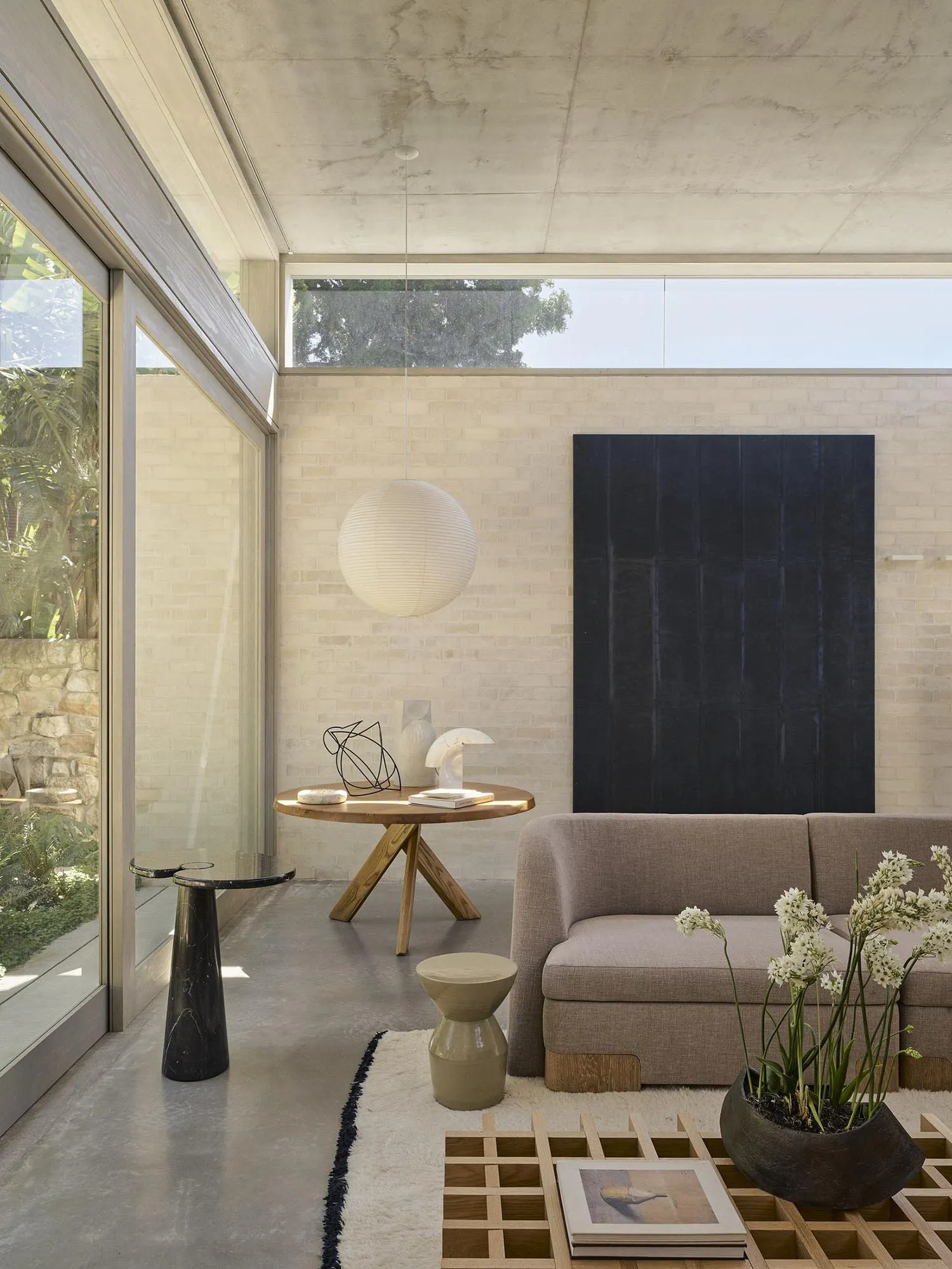 Modern minimalist living room featuring exposed concrete walls and ceiling, a beige oak-base sofa, round wooden coffee table, large black abstract painting, and a large white paper lantern pendant lamp.