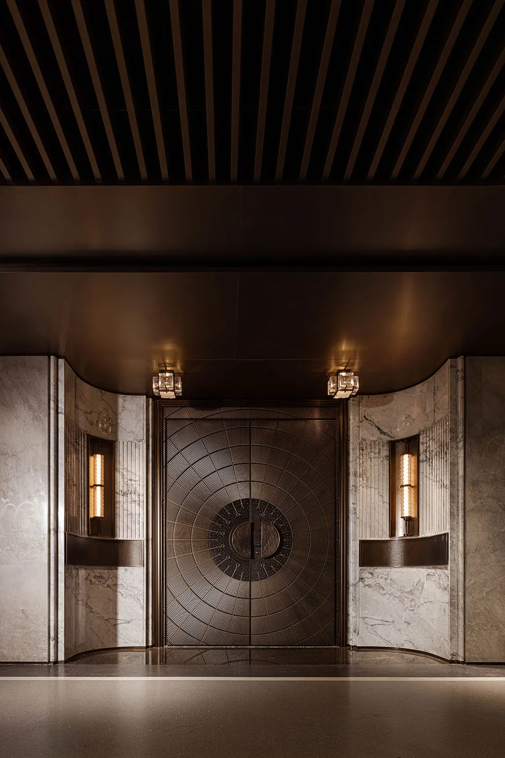 Luxurious Art Deco style entrance featuring a large bronze double door with intricate sunburst pattern, flanked by white marble walls with vertical fluting and illuminated wall sconces under a dark coffered ceiling.