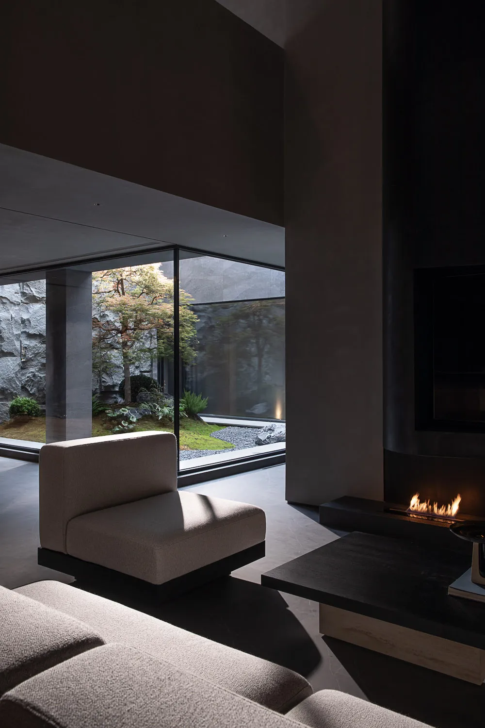 Modern minimalist living room interior featuring raw concrete walls, a built-in linear fireplace, low-profile beige fabric sofa, and a tranquil Japanese garden visible through floor-to-ceiling glass windows.