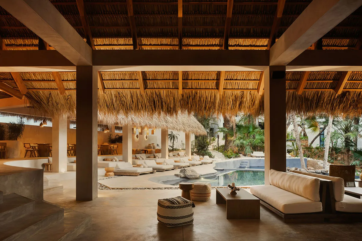 Spacious open-air lounge in a tropical villa featuring concrete pillars, thatched roof, cream-colored sofas, woven poufs, and an infinity pool overlooking lush palm trees.