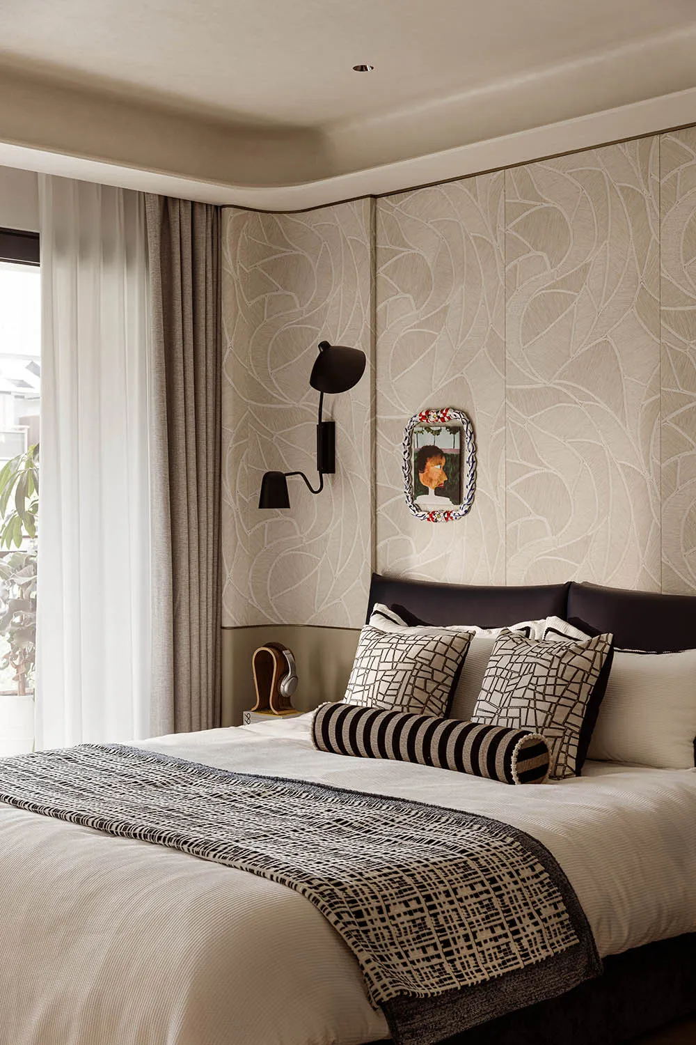 Modern bedroom interior with beige leaf-patterned textured wall panels, dark leather headboard, black wall sconce, and bed dressed in white linen with black and beige geometric patterned pillows and throw.